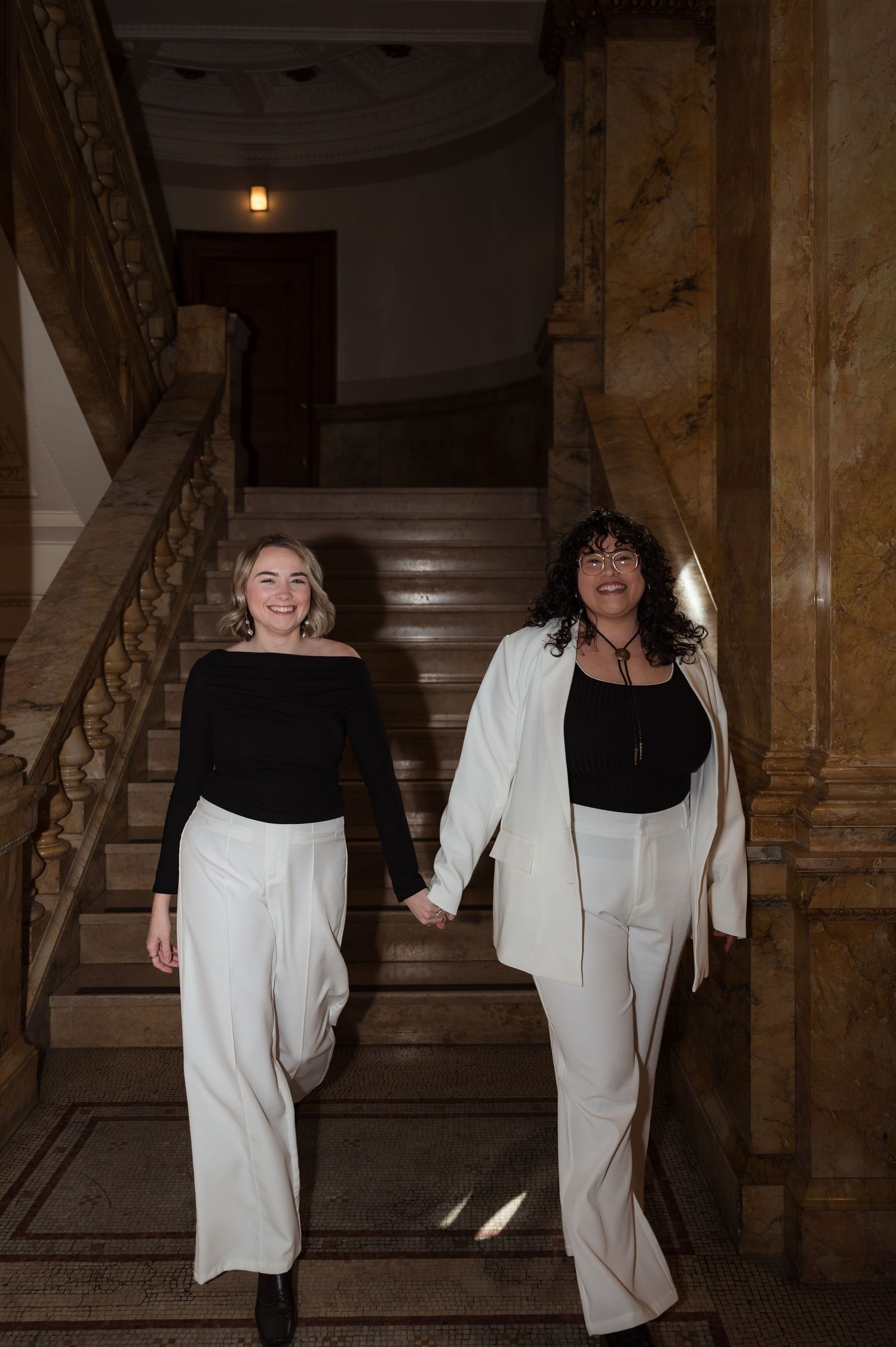 Queer couple in matching black and white outfits smiling while descending marble staircase.