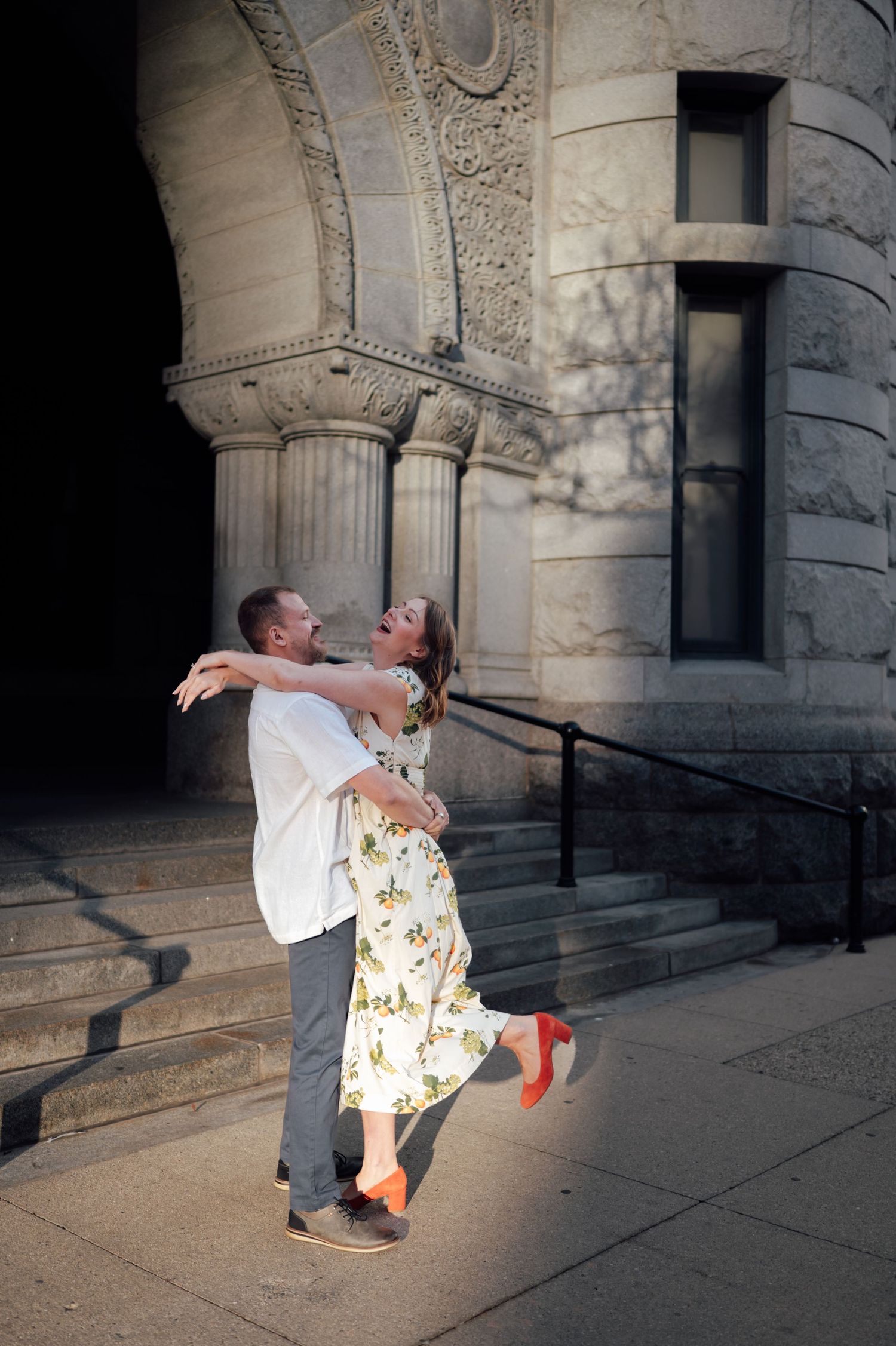 Man joyfully lifting woman in floral dress outside a historic stone building with ornate architectural details.