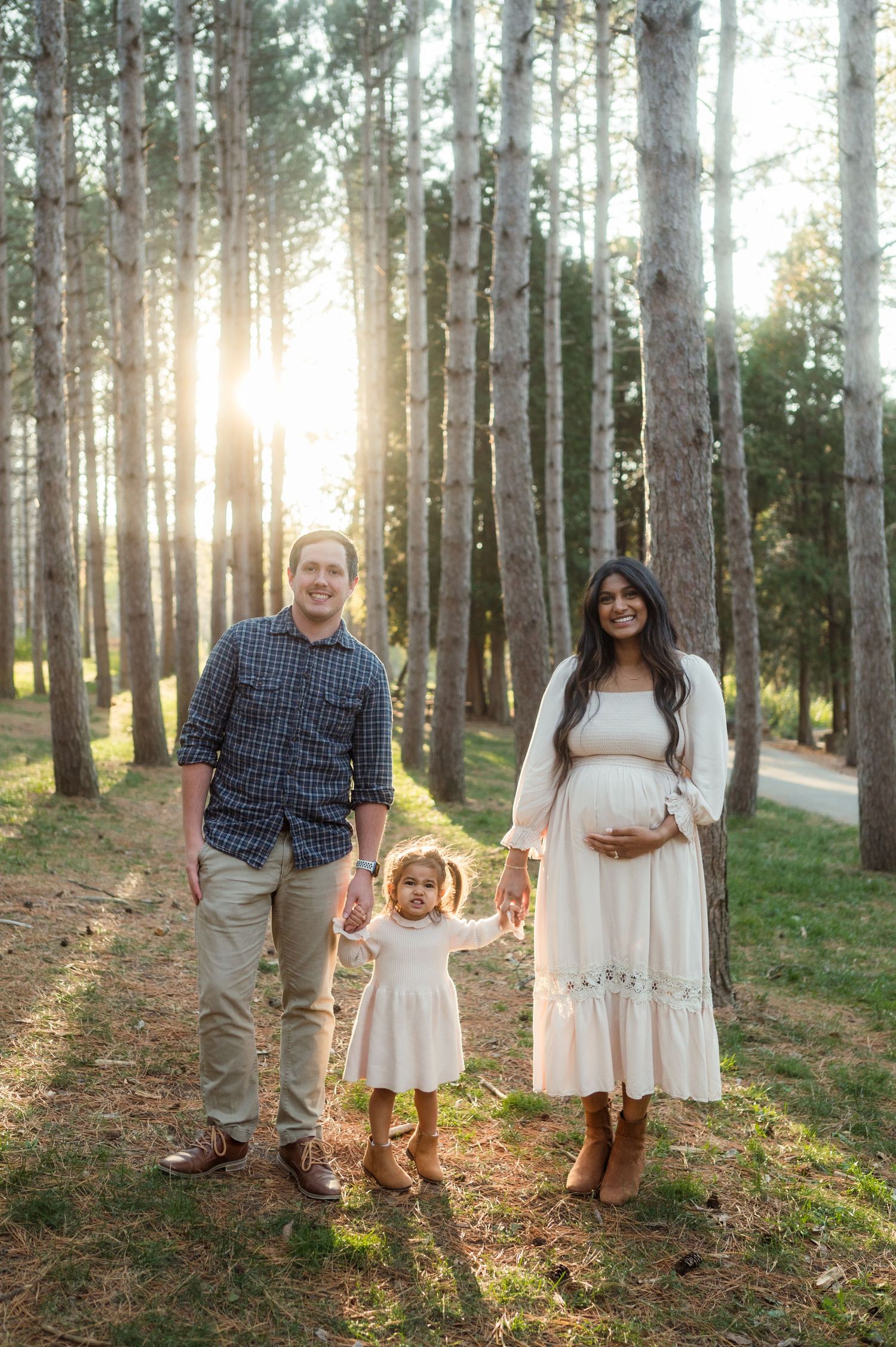 Expectant family of three holding hands in sunlit pine forest, mother in white maternity dress.