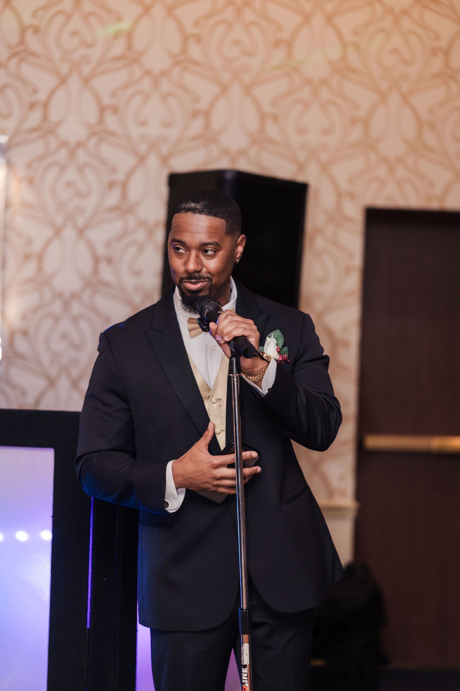 A speaker in a dark suit holds a microphone while speaking at an event with purple lighting.