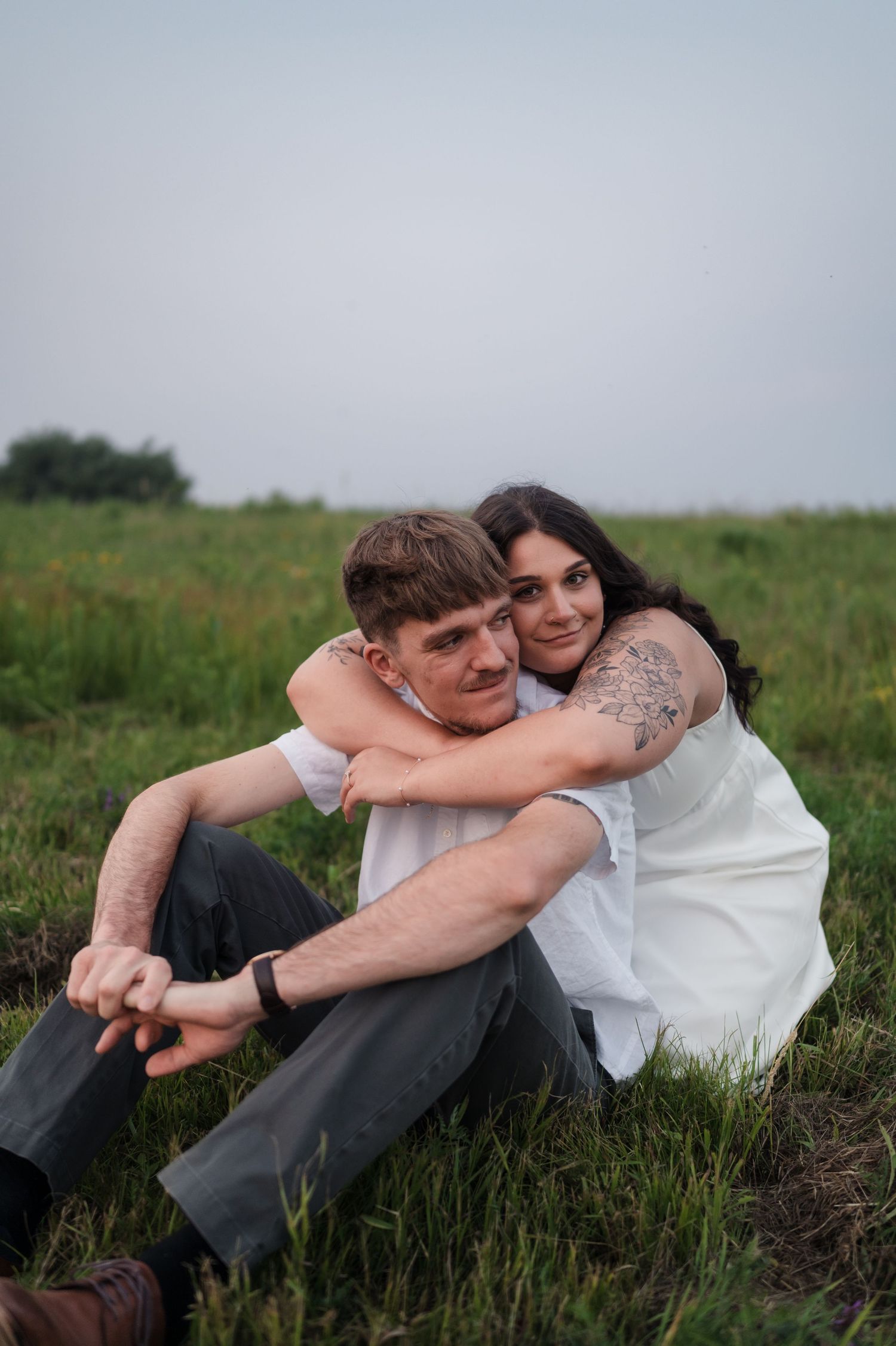 A couple embraces while sitting together in a grassy field on an overcast day.