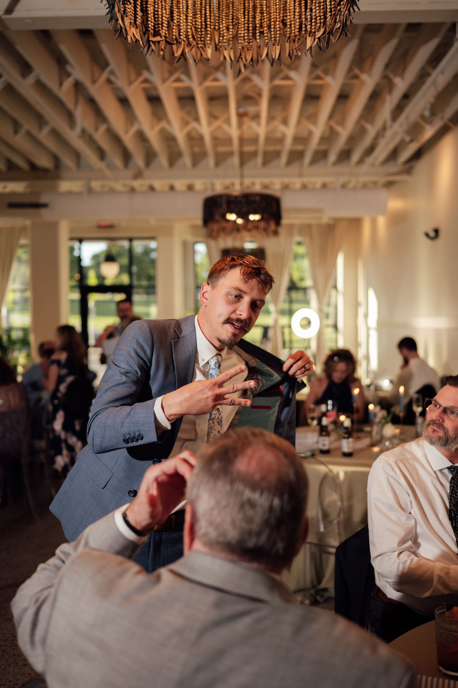 A man in a blue suit holds his tie while speaking to guests at an elegant wedding reception venue.