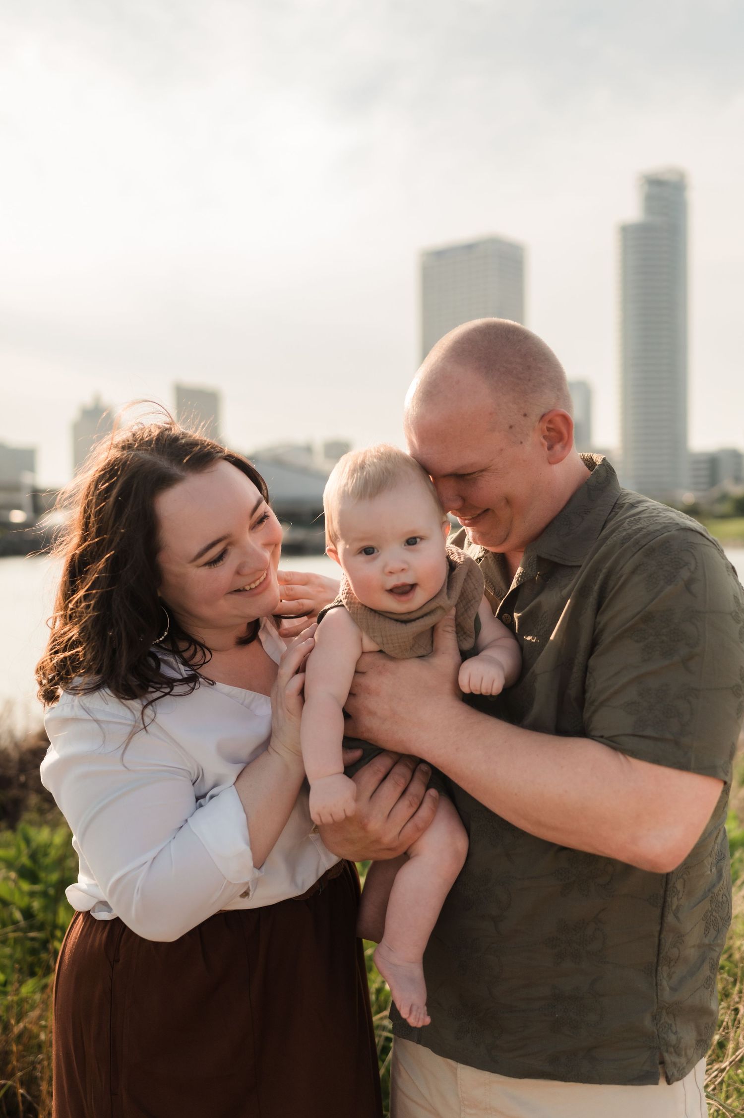 Young couple holding their smiling baby outdoors with a city skyline in the background at sunset.