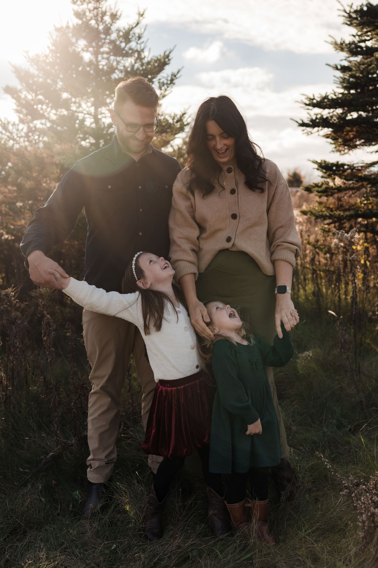 Happy family of four with two young girls laughing in a sunlit pine tree field.