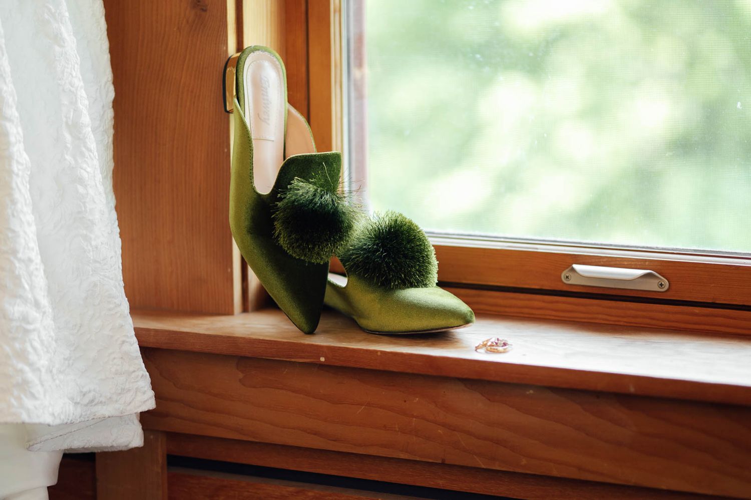 Green satin wedding heels with fur pom-pom detail displayed on a wooden windowsill.