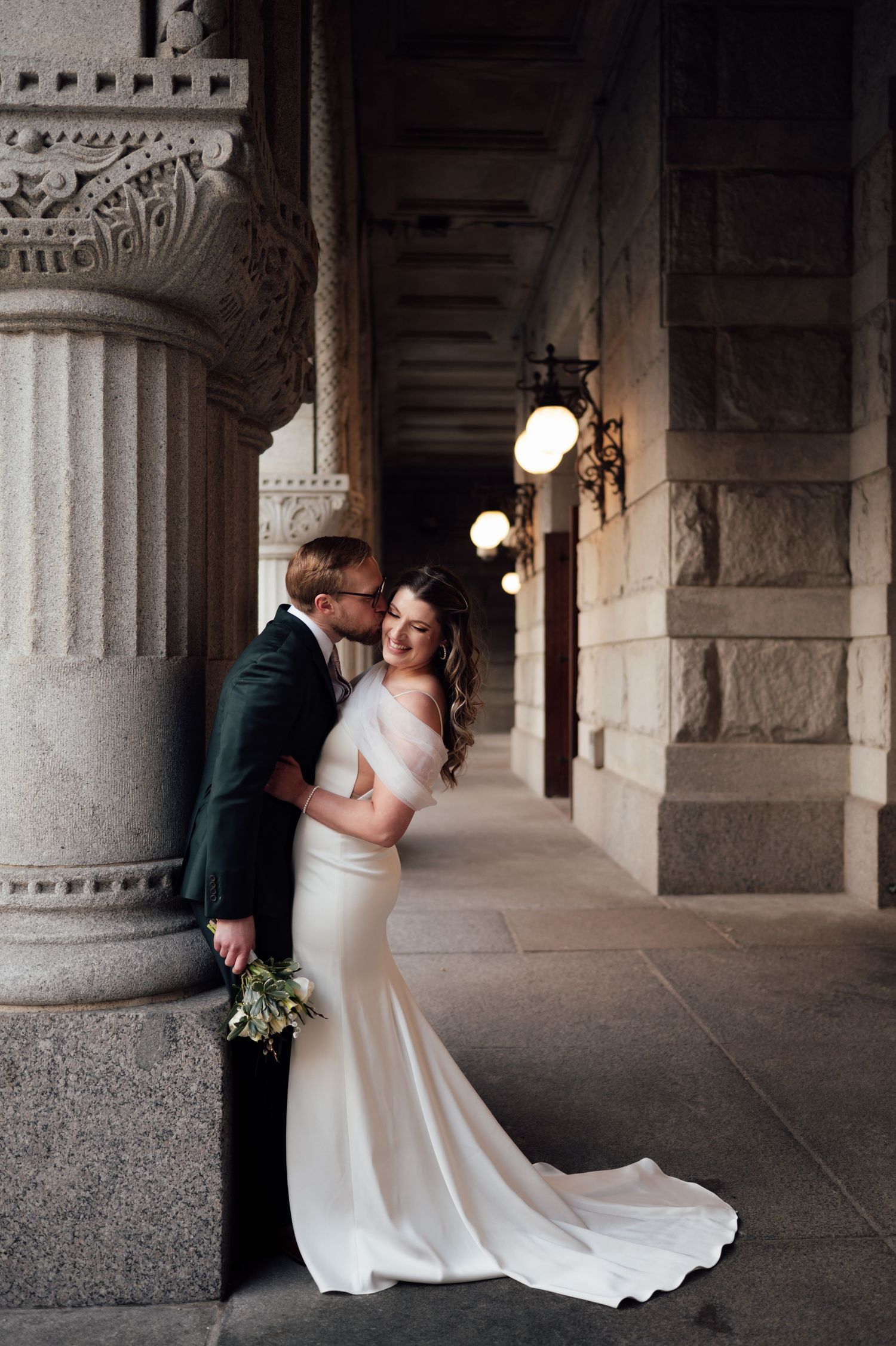 A bride in a fitted white gown leans against a stone column as her groom kisses her cheek.
