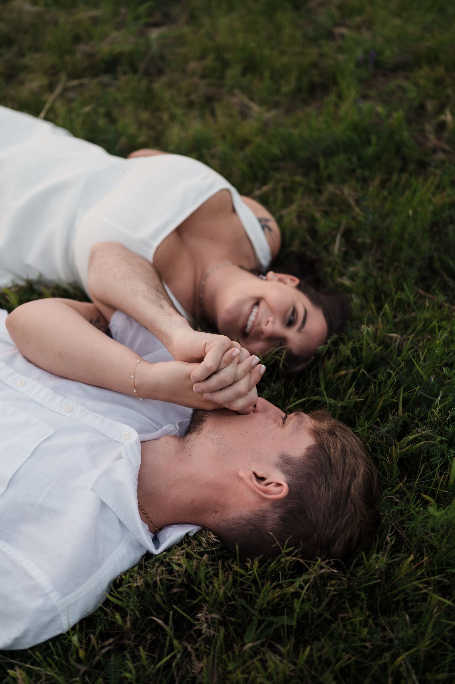 Happy couple lying in green grass holding hands and laughing together during engagement shoot.