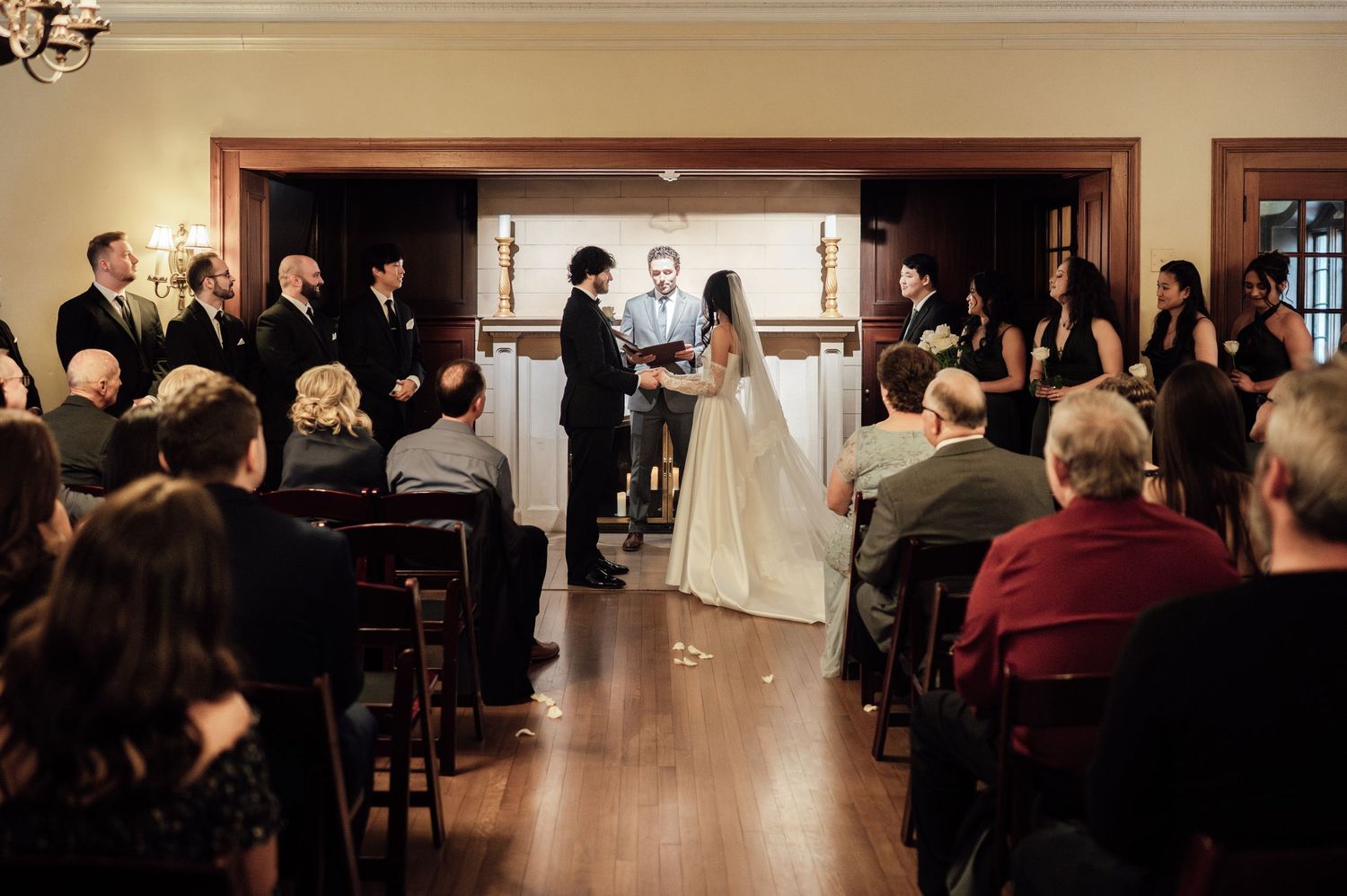 Indoor wedding ceremony with bride and groom exchanging vows before officiant, guests seated in wooden pews, elegant venue.