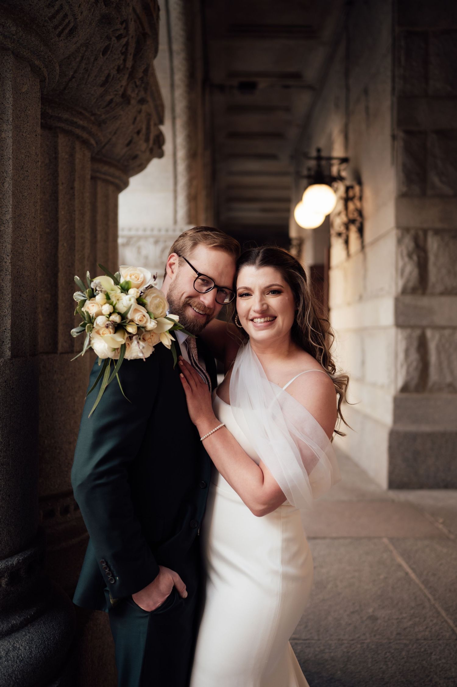 Smiling bride and groom pose together in a grand stone hallway, bride in white gown with veil holding bouquet.