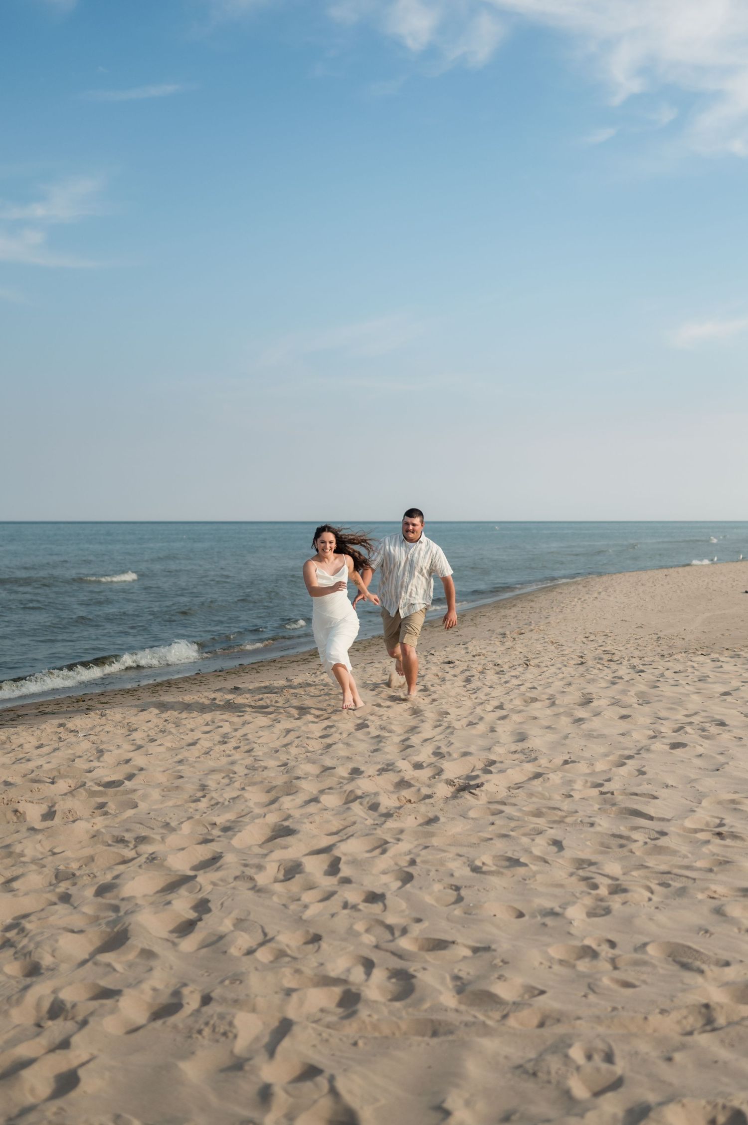 Couple walking along a sandy beach shoreline while holding hands on a sunny day with ocean waves in the background.