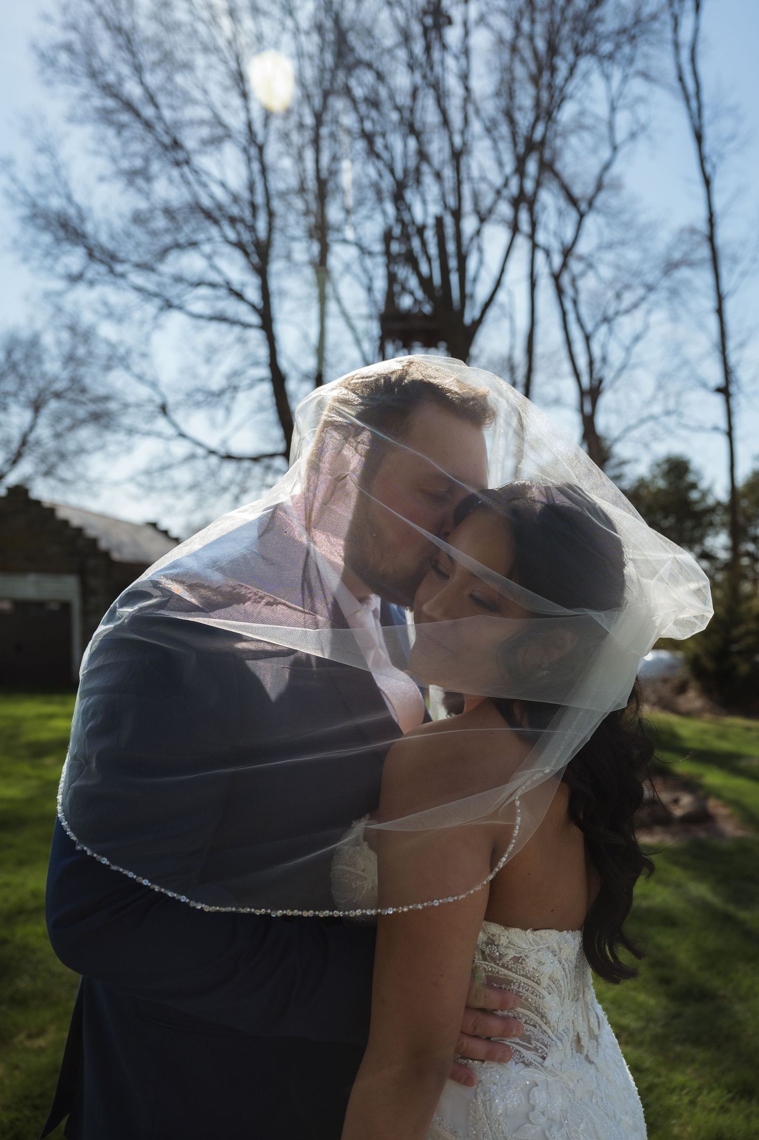 A romantic wedding moment with a couple sharing a kiss under a flowing veil outdoors on a sunny day.