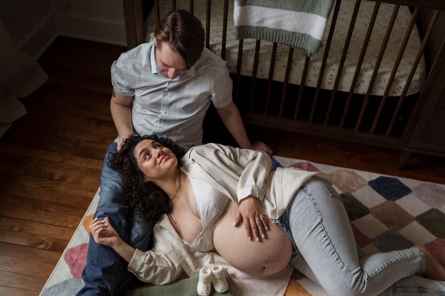 Couple sharing tender moment on nursery floor, pregnant woman resting with partner by crib.