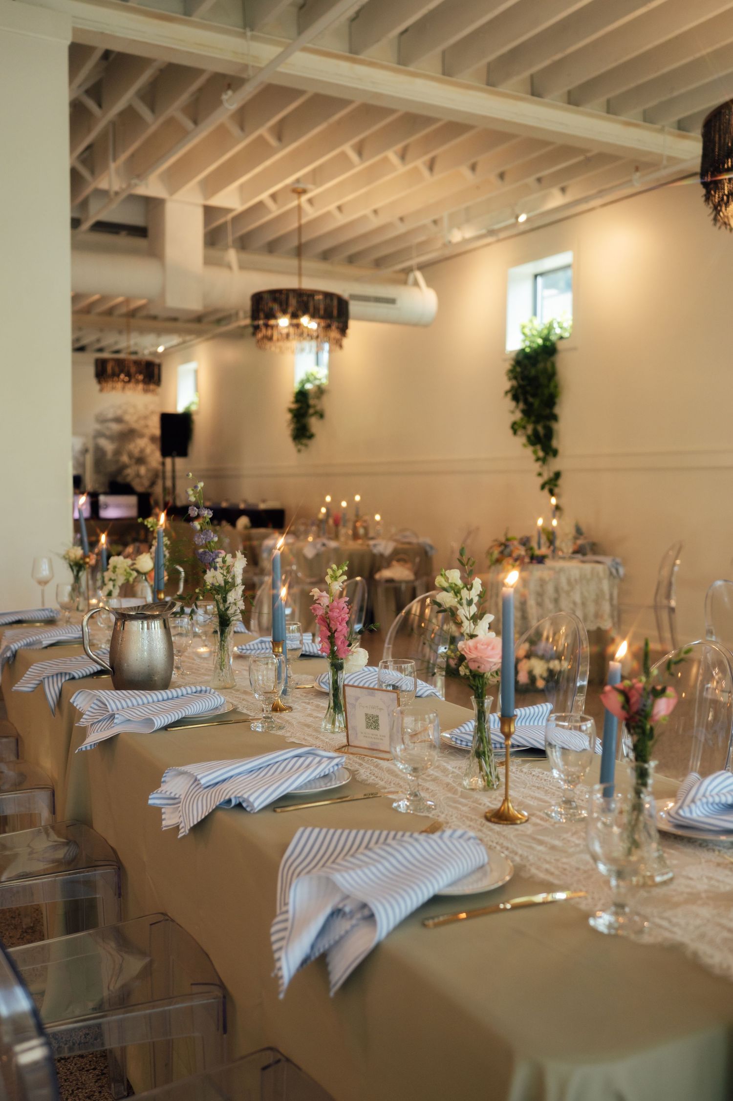 Elegant wedding reception table set with floral centerpieces, candles, striped napkins, and glassware in a bright venue.