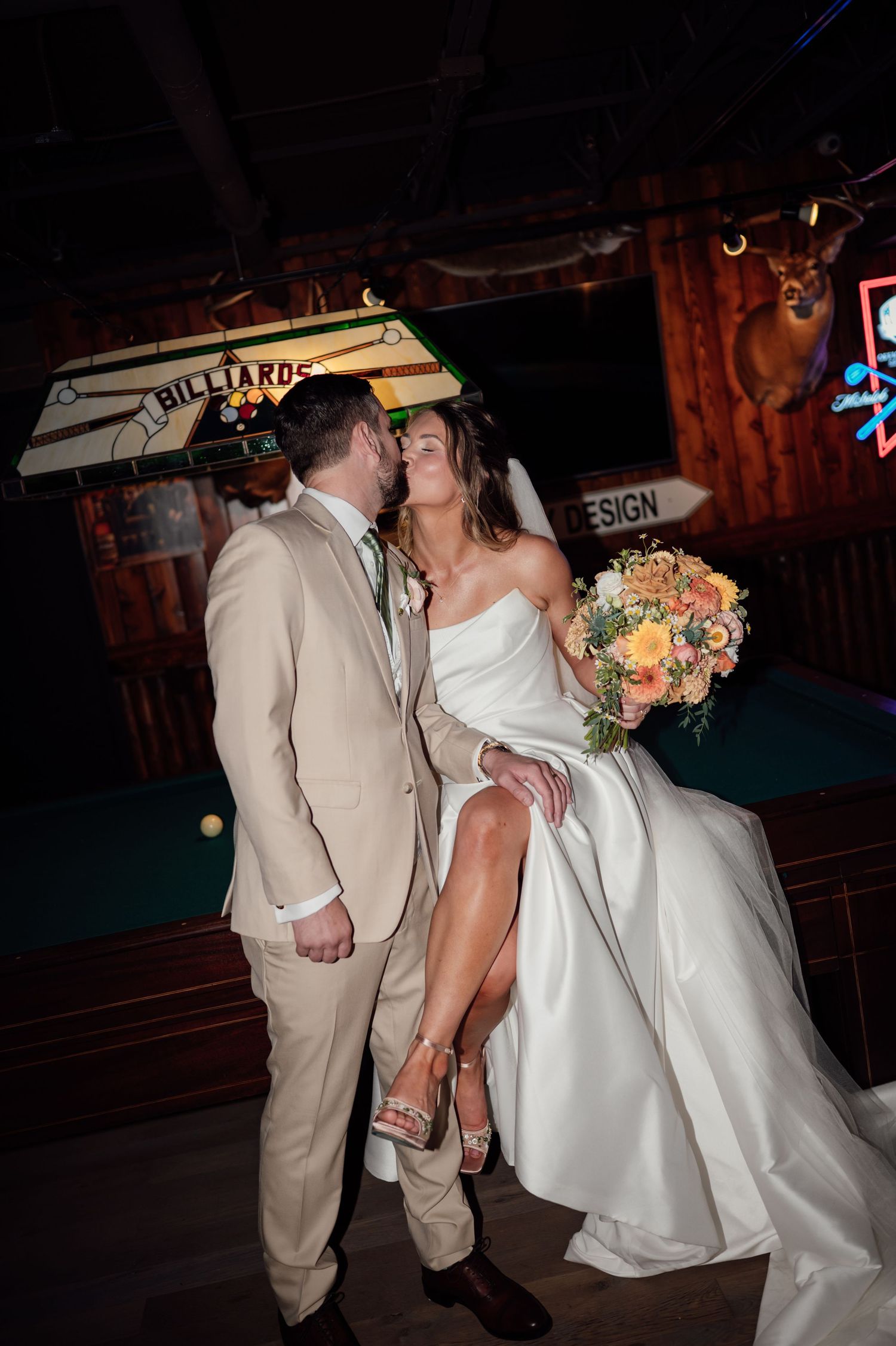 Bride and groom share a kiss near a billiards table, she holds a colorful bouquet, wearing a white gown with a slit.