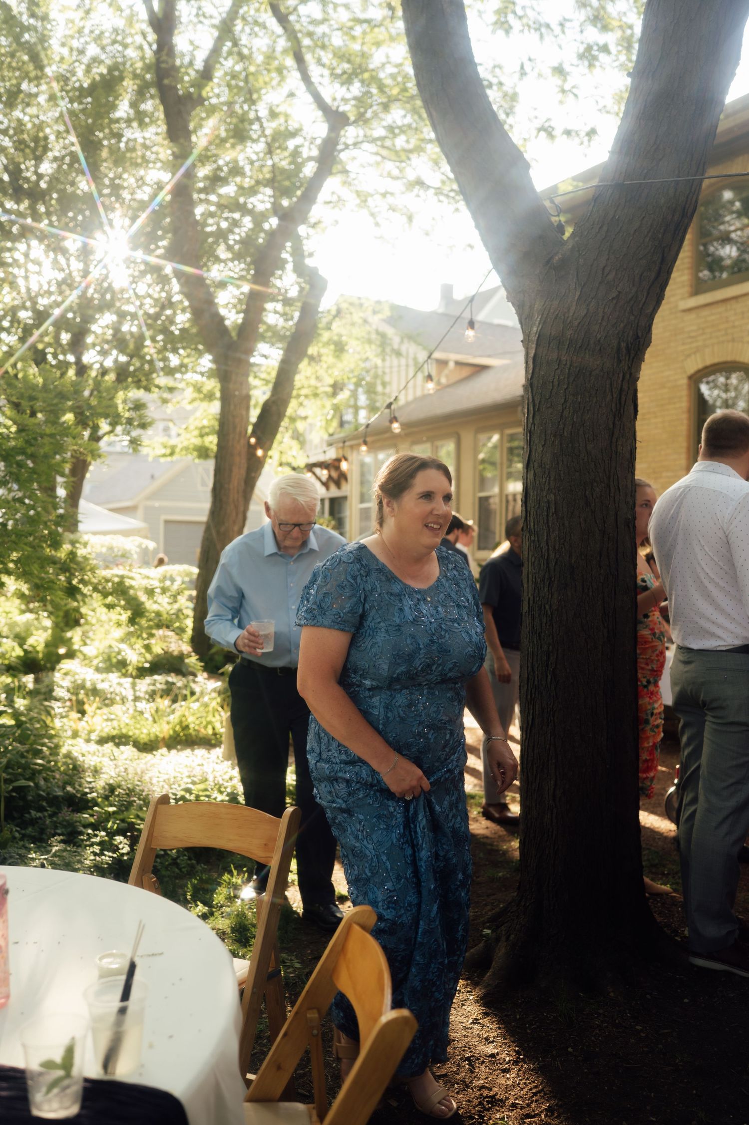 Woman in blue floral dress socializing at an outdoor garden party with guests and trees in sunlight.