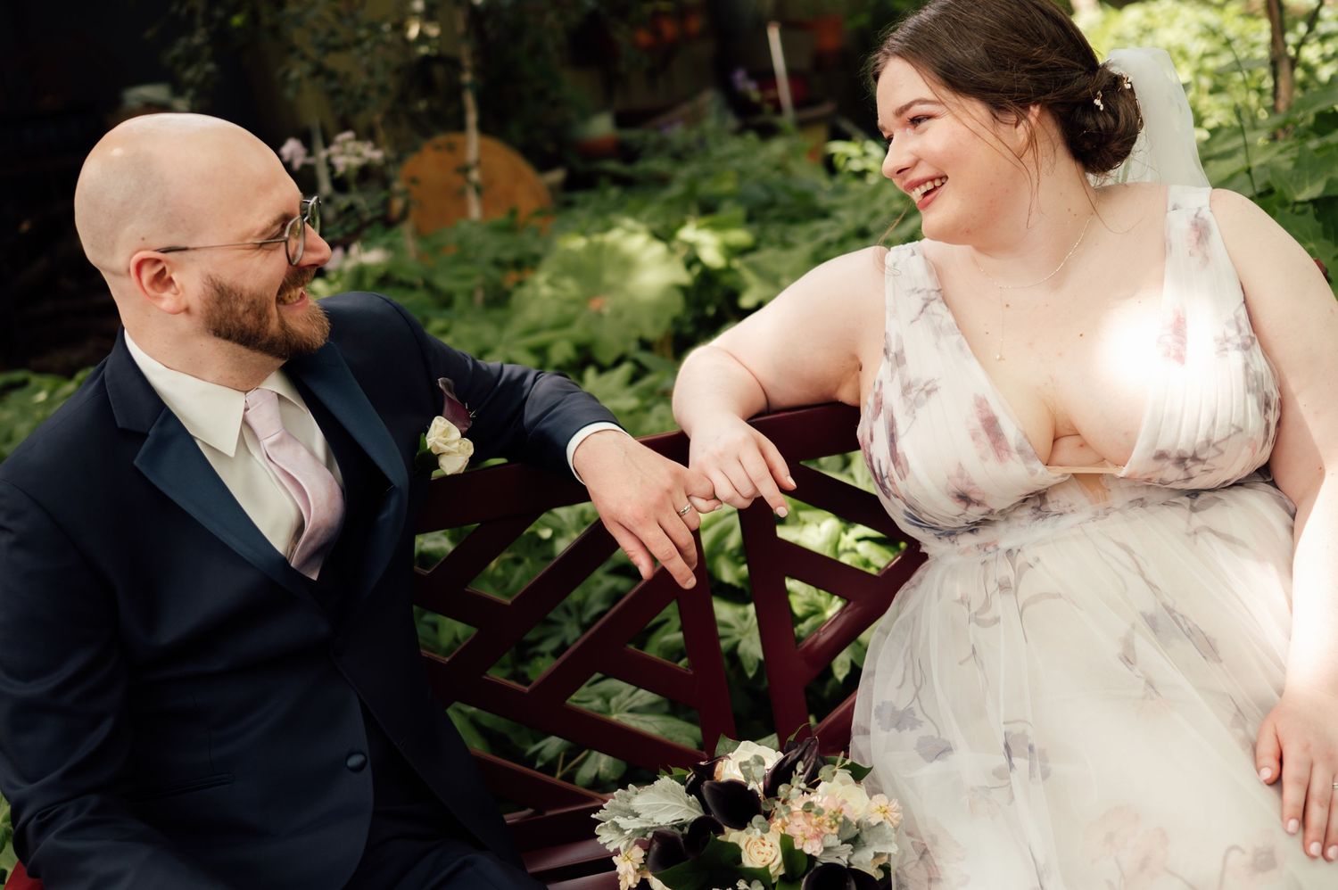 A bald groom in a navy suit and a bride in a floral gown share a joyful moment on a garden bench.