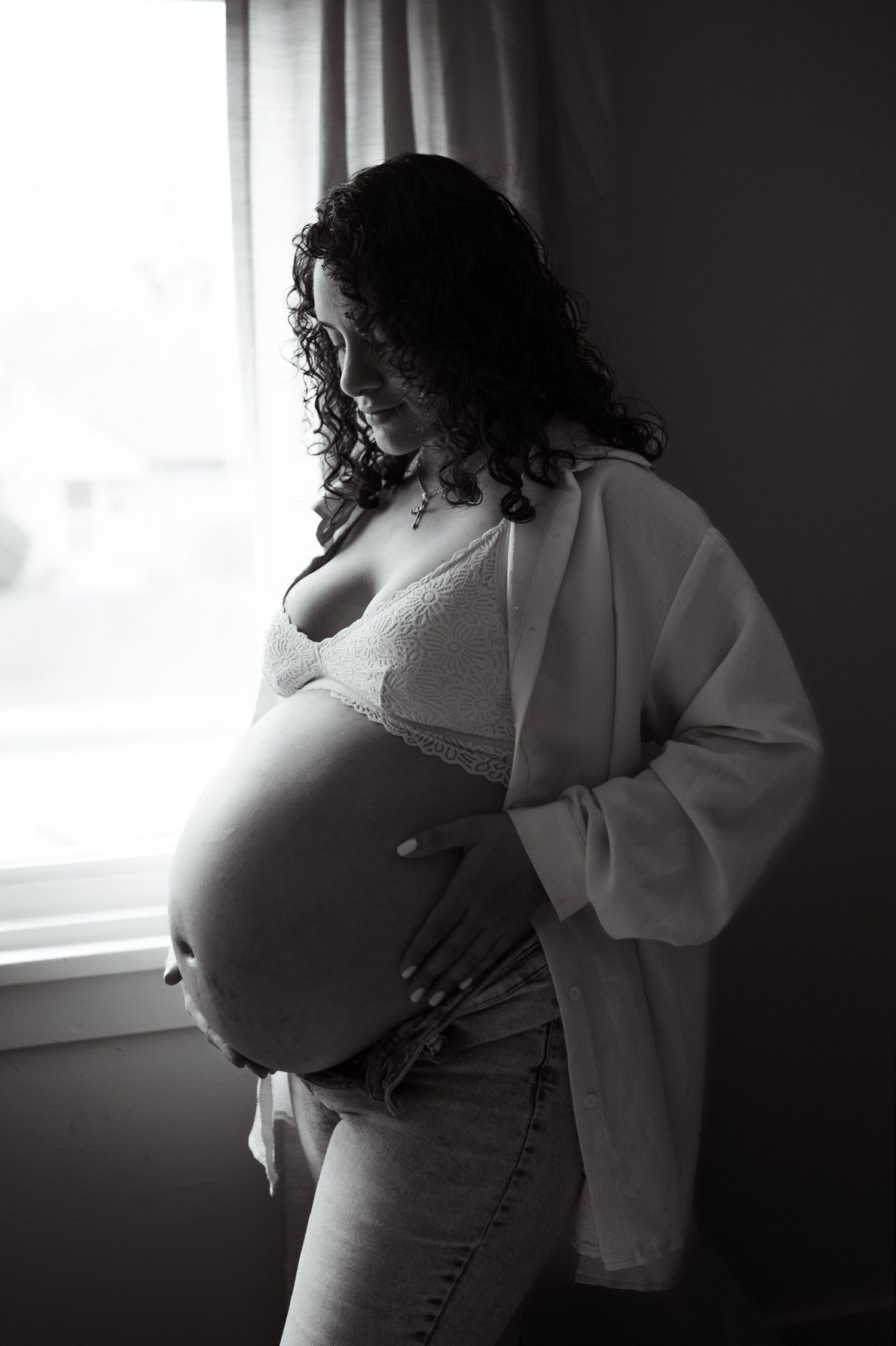 Black and white portrait of an expectant mother by window light wearing white cardigan and jeans.
