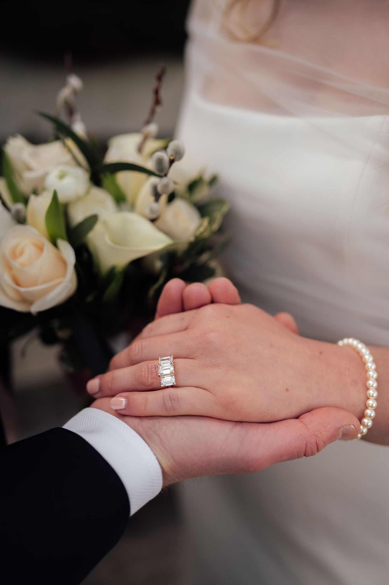 Bride and groom holding hands showing diamond engagement ring and pearl bracelet with white rose bouquet.