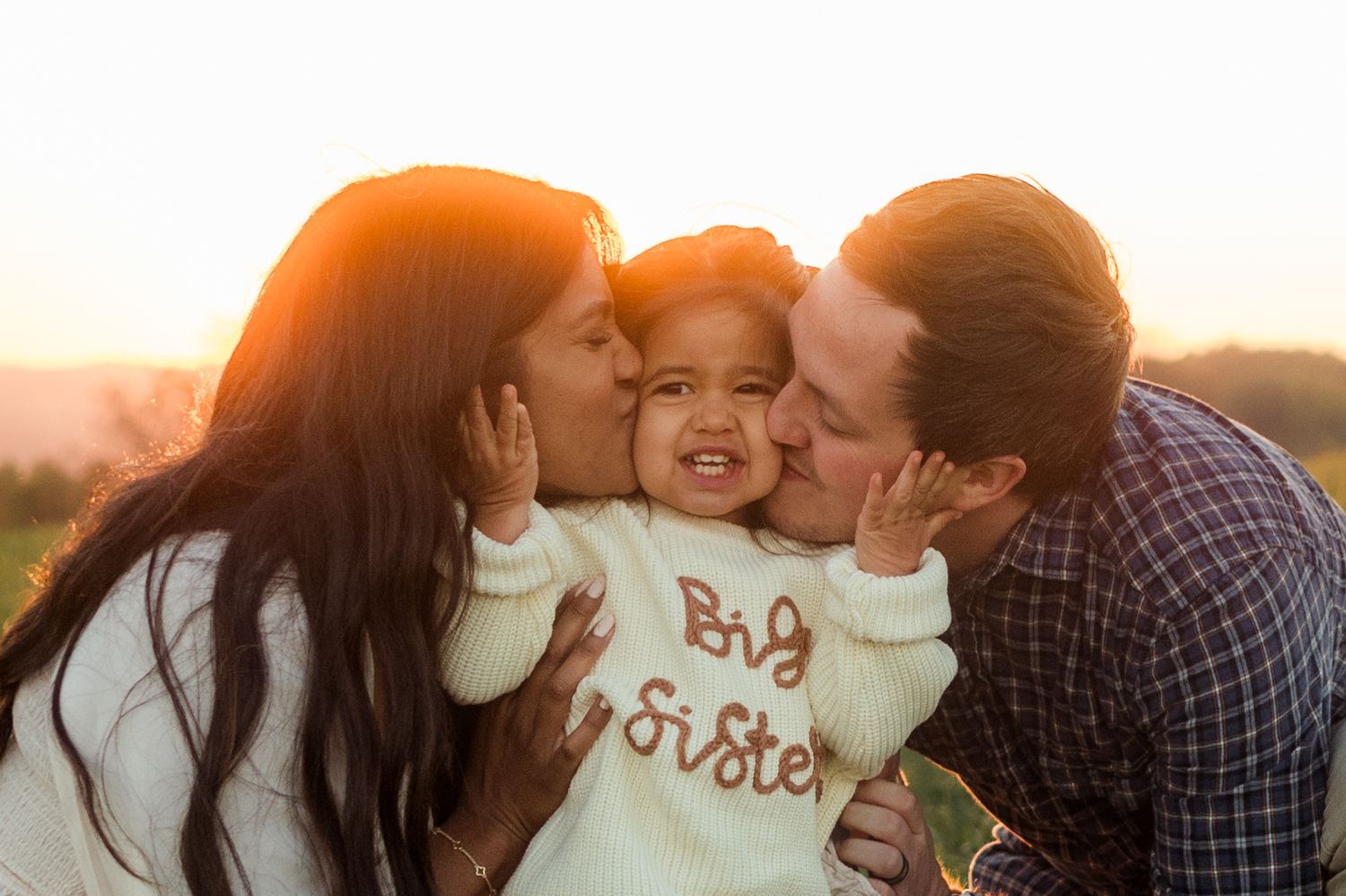 Parents kissing toddler wearing Big Sister sweater during golden hour pregnancy announcement photo.