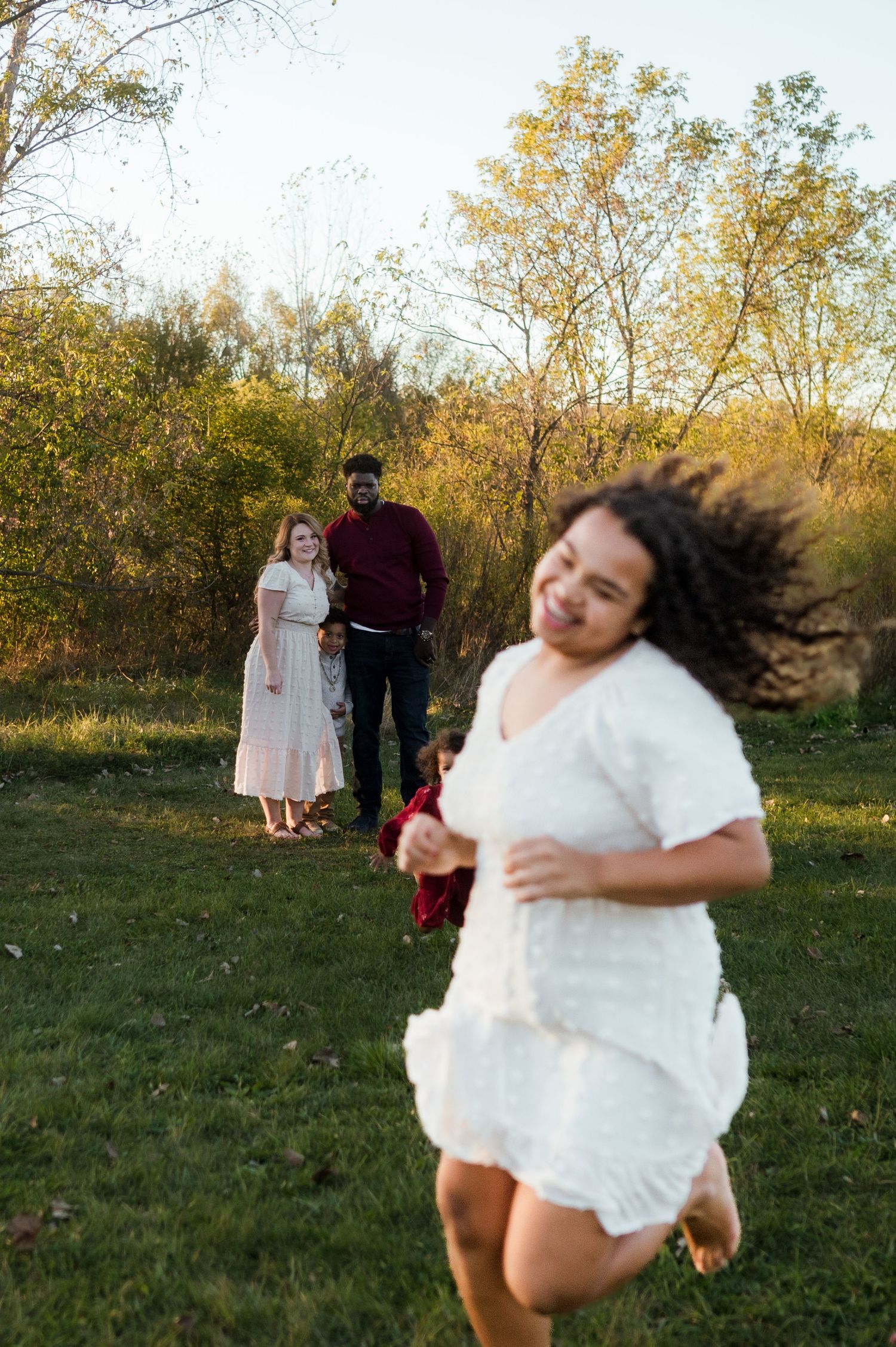A joyful girl in white spins outdoors while her interracial family smiles warmly in an autumn park setting.