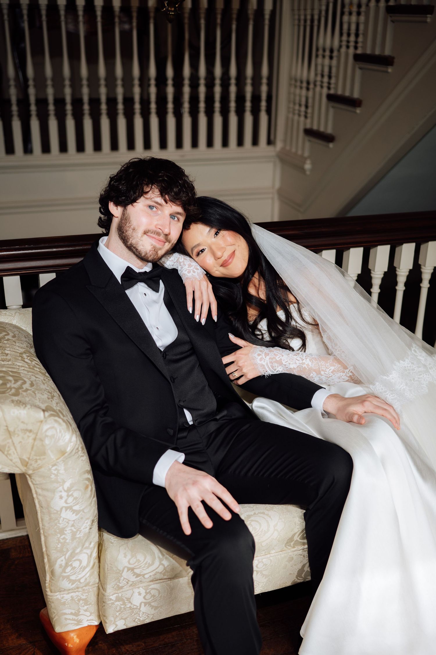 Newlywed couple in elegant wedding attire sits together on ornate sofa near grand staircase.