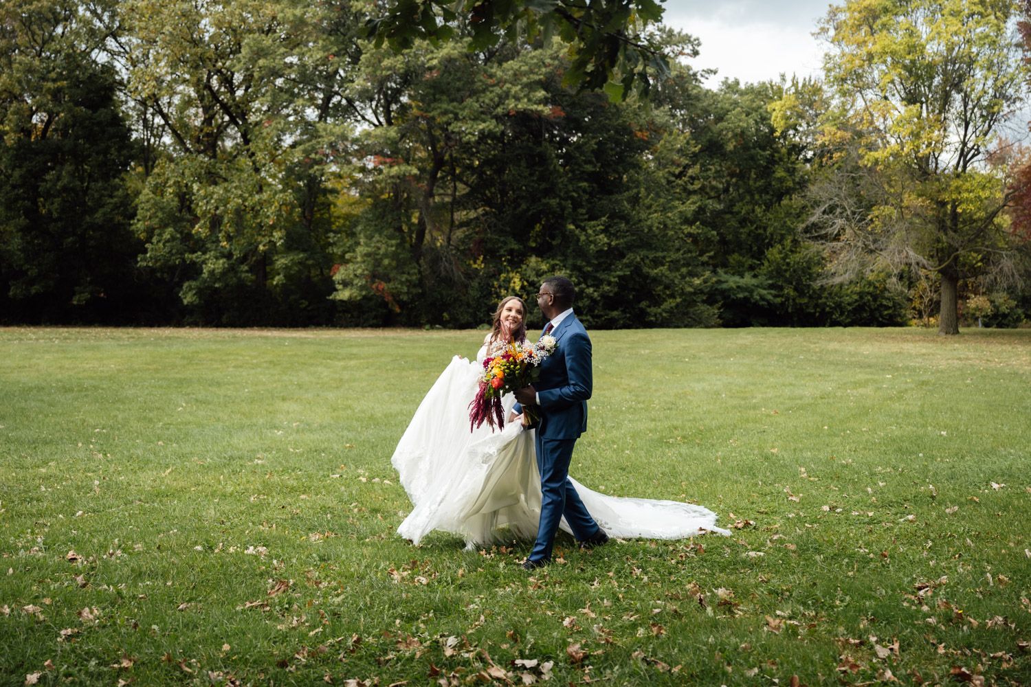 Bride and groom dance together on green lawn surrounded by autumn trees in outdoor wedding portrait.