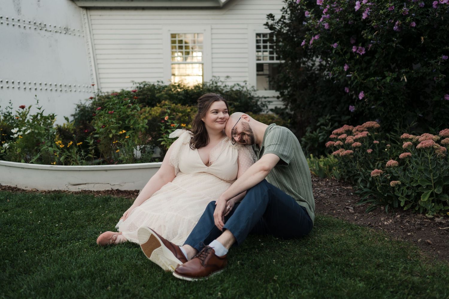 Couple relaxing on grass leaning against each other near blooming garden outside a white historic building.