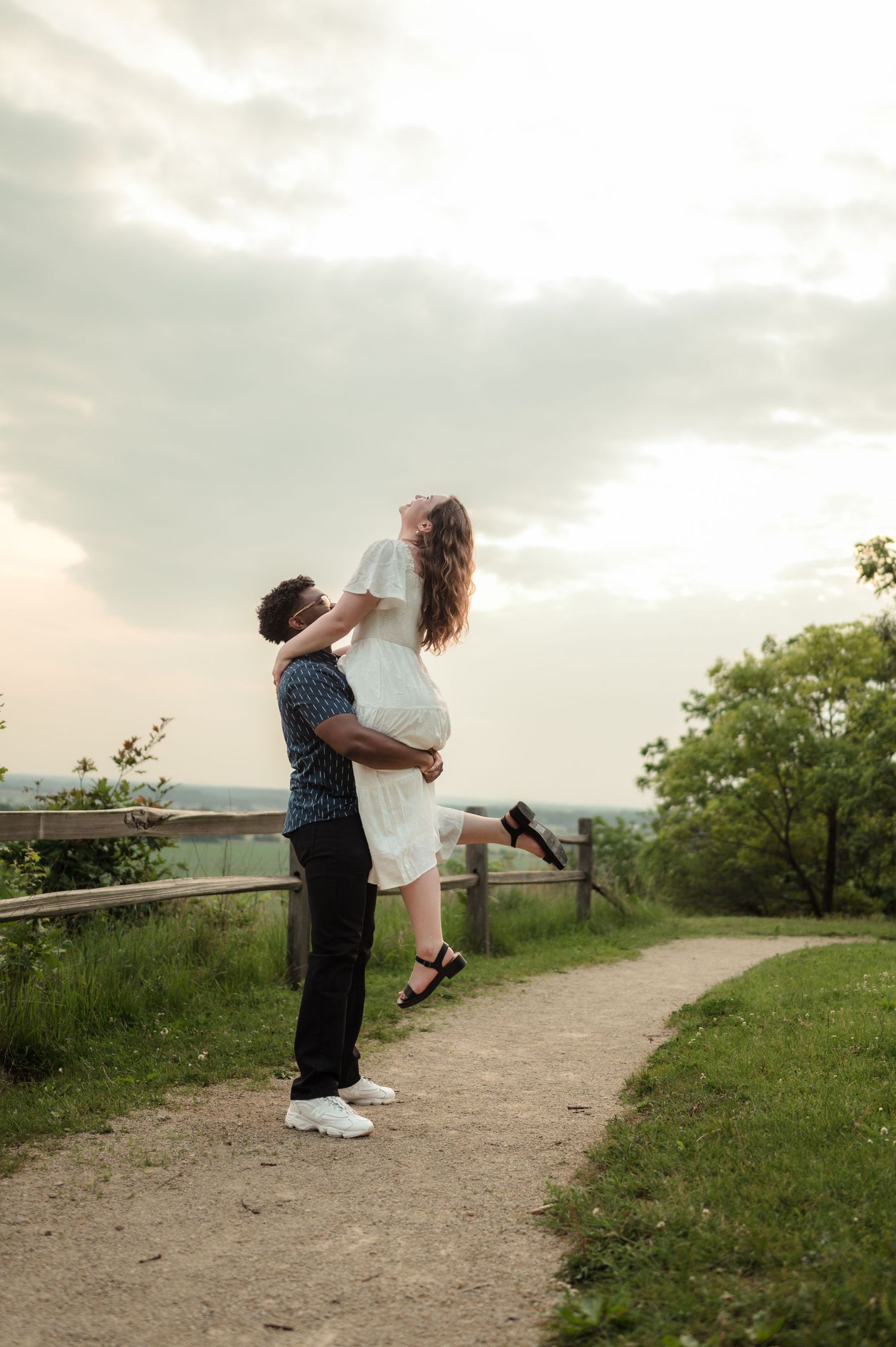 Man lifting woman in white dress on a rustic garden path with wooden fence and lush greenery.