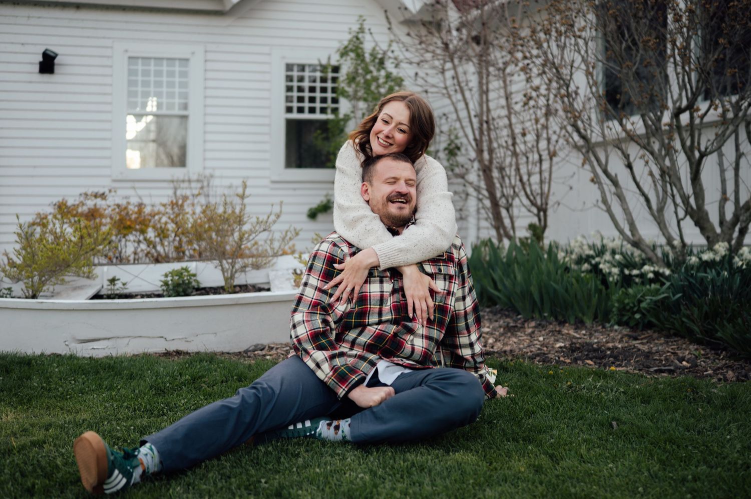 Couple laughing together on green grass in front of a white house during an outdoor engagement photo session.
