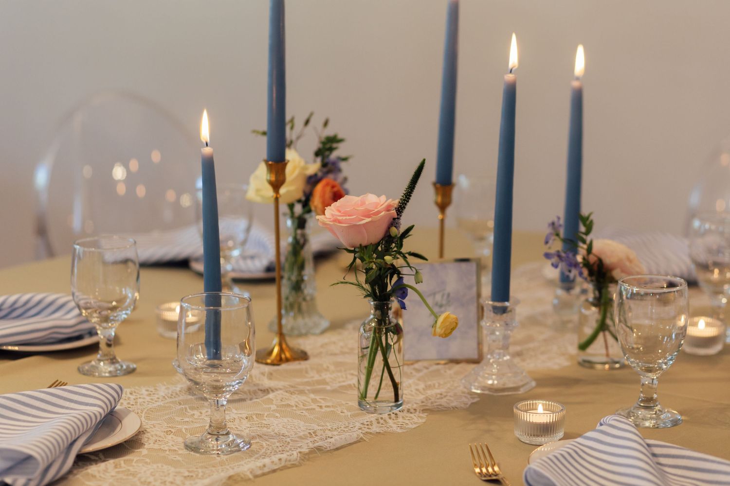Elegant wedding table setting with blue taper candles, pink roses, striped napkins, and gold cutlery on a lace runner.