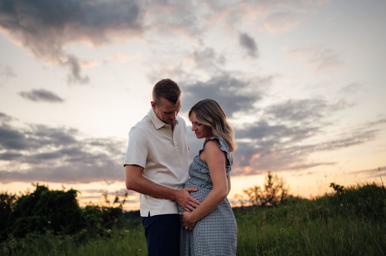 Couple embracing at sunset in a field, woman showing baby bump, romantic maternity photo with dramatic cloudy sky.