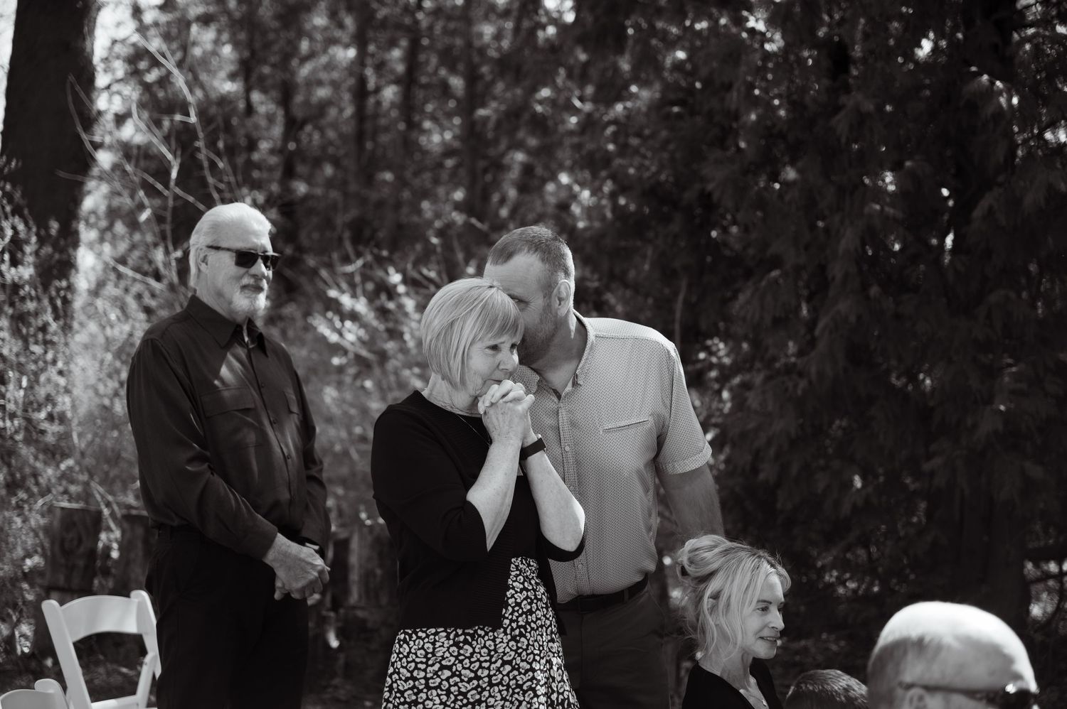 An emotional black and white candid moment captured during an outdoor gathering with guests looking on.