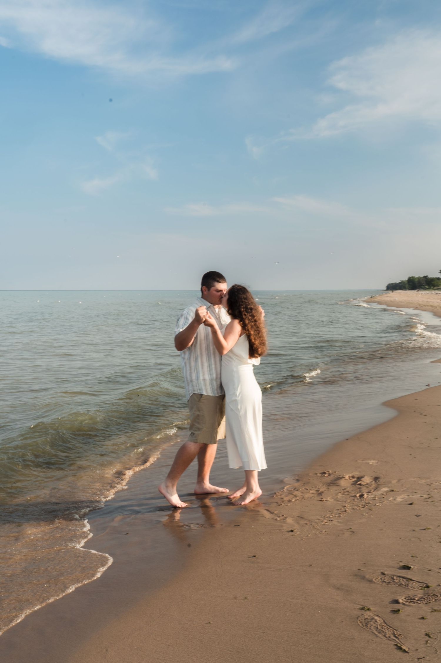 A couple dances together barefoot on a sandy beach at sunset with gentle waves lapping at the shore.