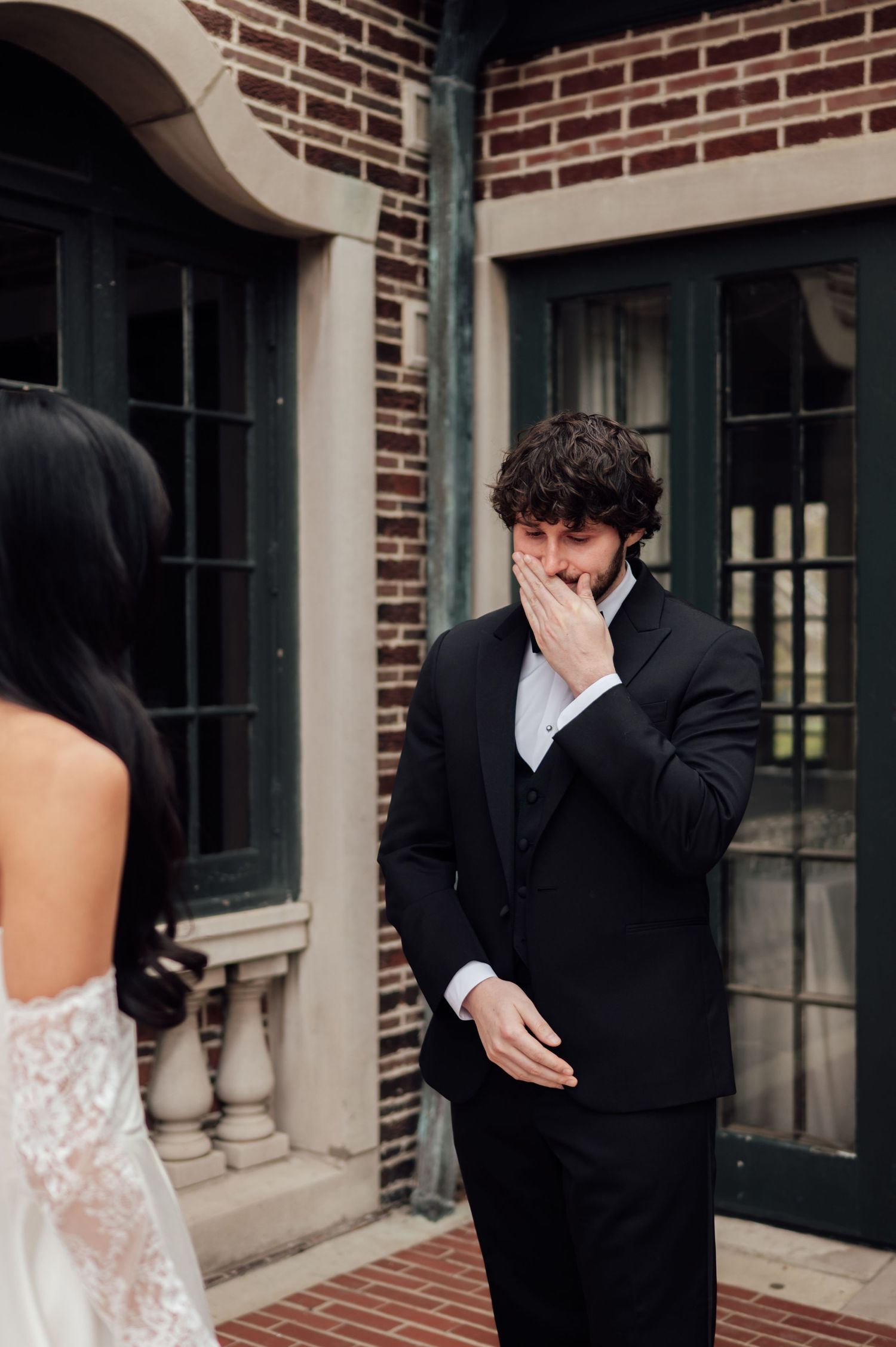 Emotional groom in black suit covering his mouth during first look moment with bride in lace wedding dress outside brick venue.