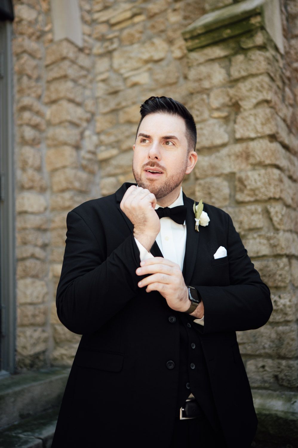 Groom in black tuxedo with bow tie and boutonniere adjusting cufflinks outside stone building.