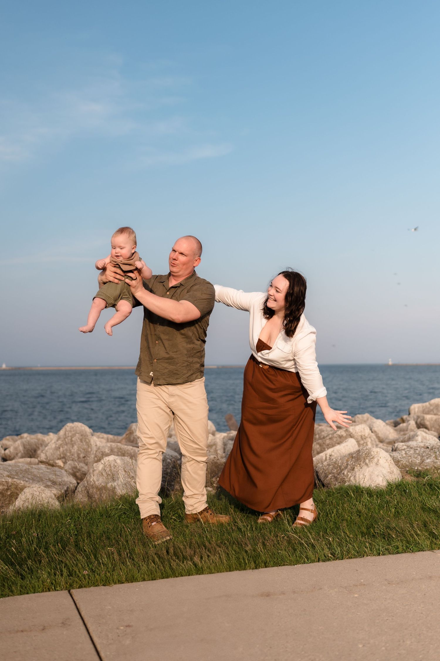 Smiling parents holding baby near rocky lakeside shore under a bright blue sky.