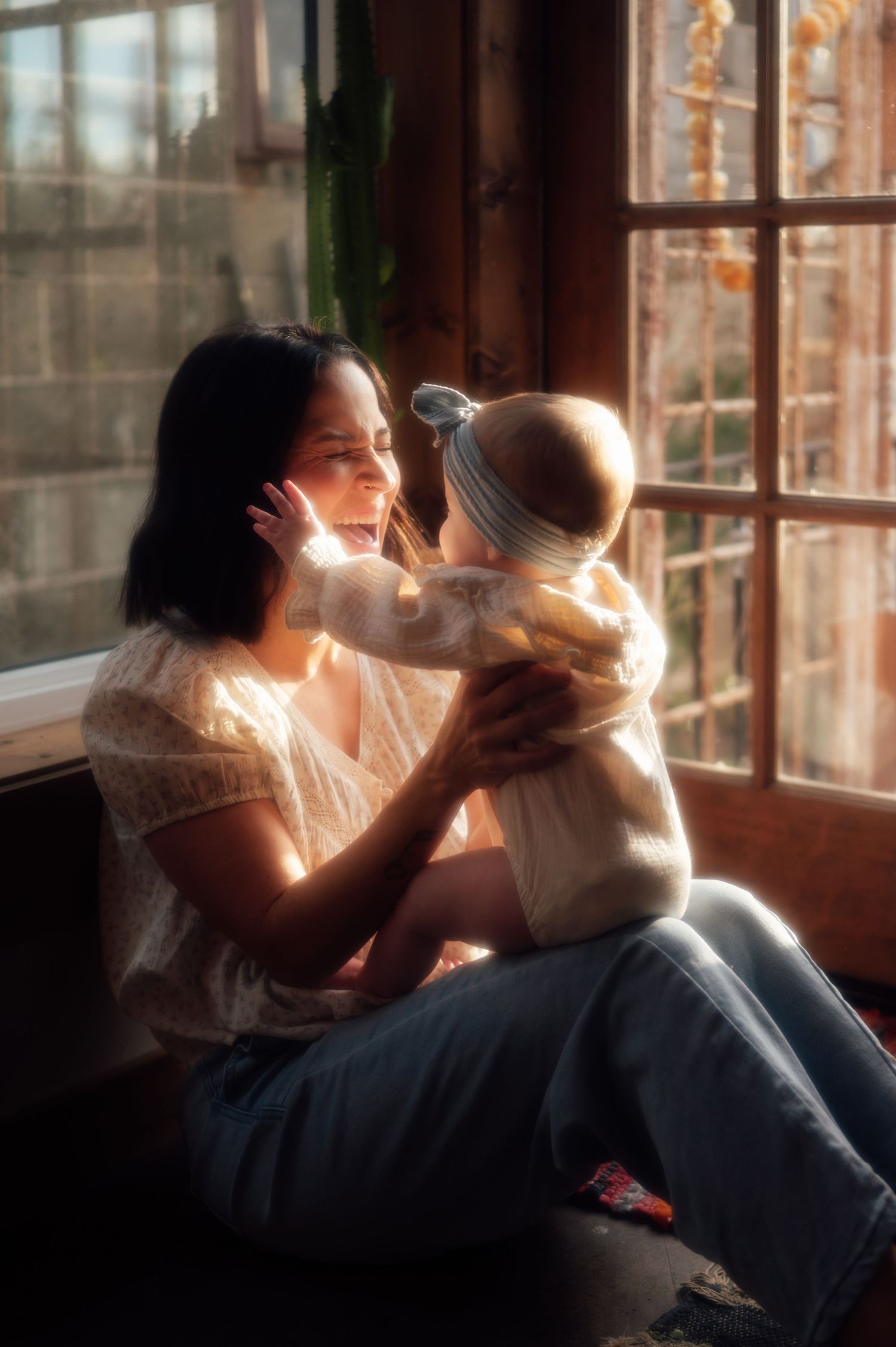 A joyful mother laughs as her baby reaches for her face while sitting by a sunlit rustic window.