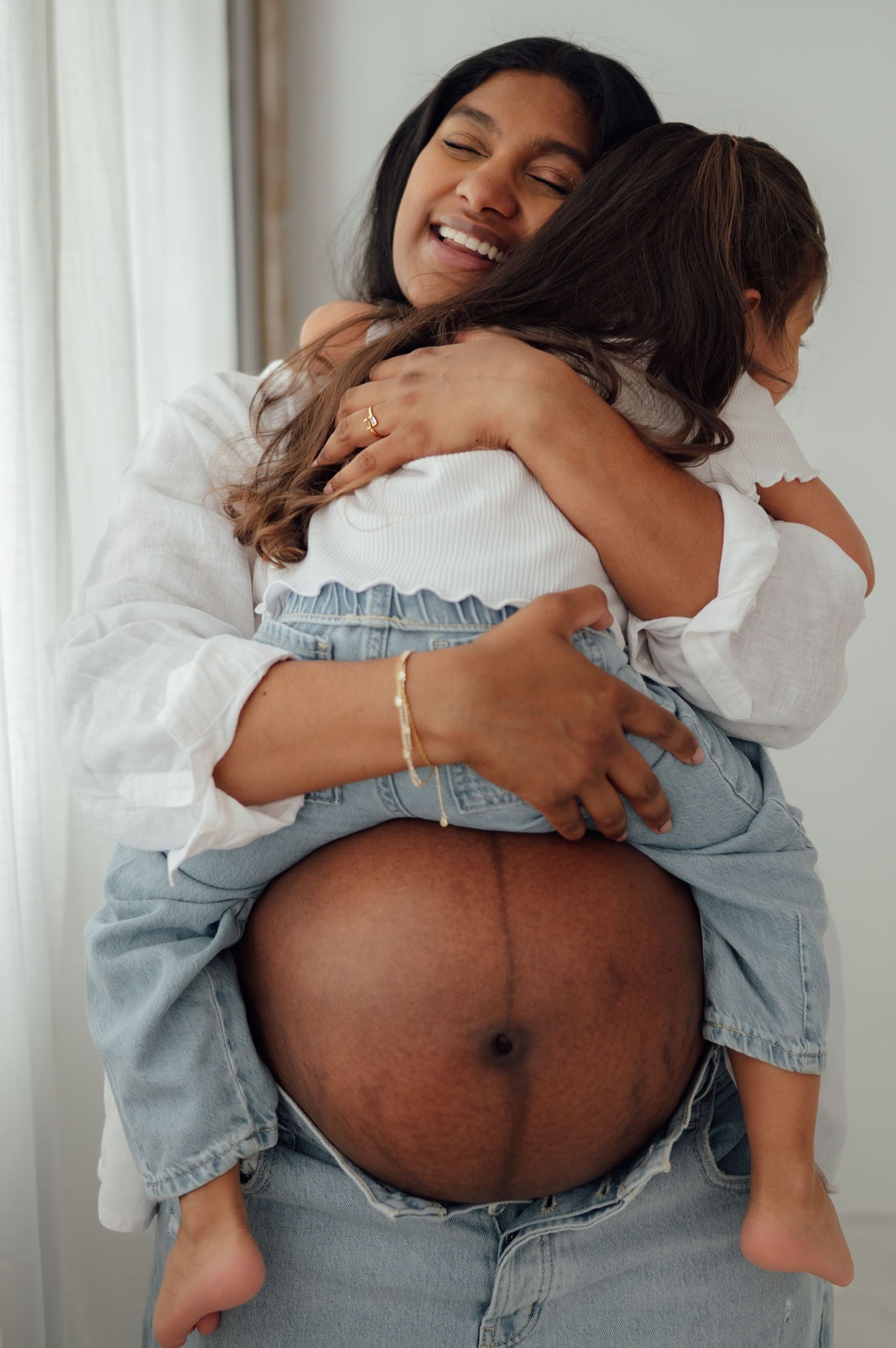 Smiling pregnant woman hugging child while showing bare baby bump in white shirt and jeans.