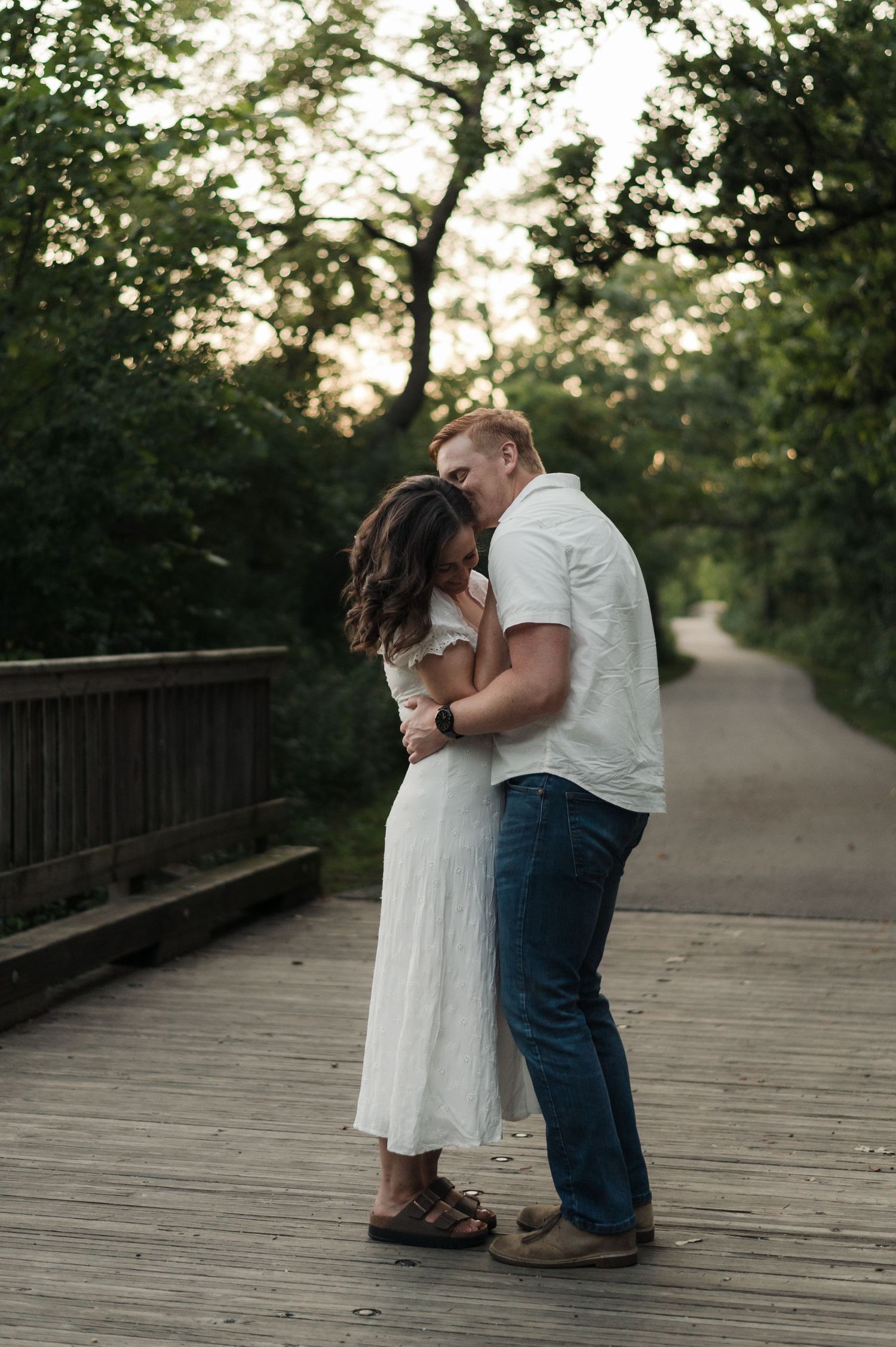 Couple shares a tender kiss on a wooden bridge path surrounded by lush green trees at dusk.