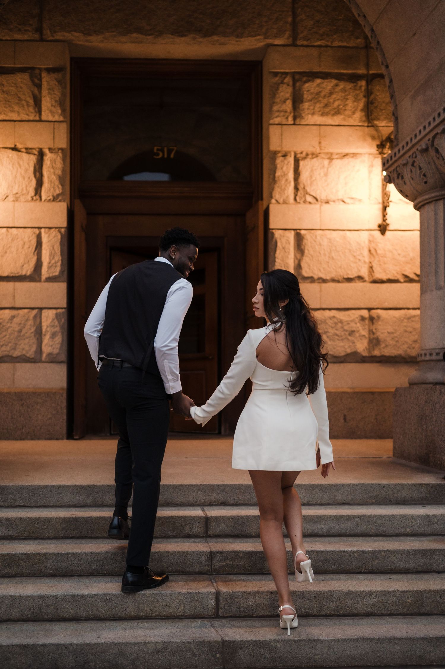 Elegant couple holding hands walking up stone steps toward a classic building entrance at dusk.