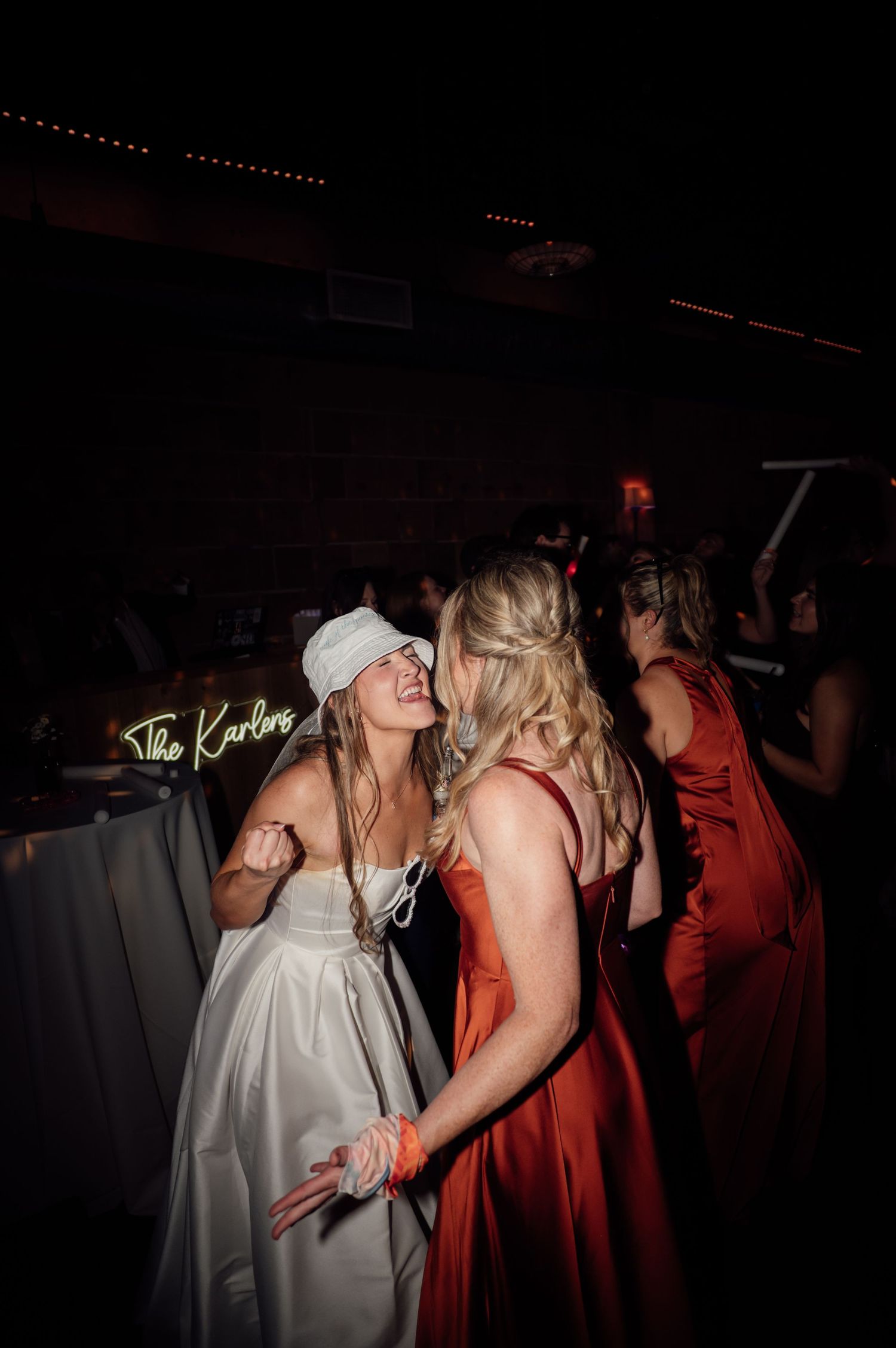 Bride in white dress and blindfold dancing with guest in orange gown at wedding reception with neon sign.