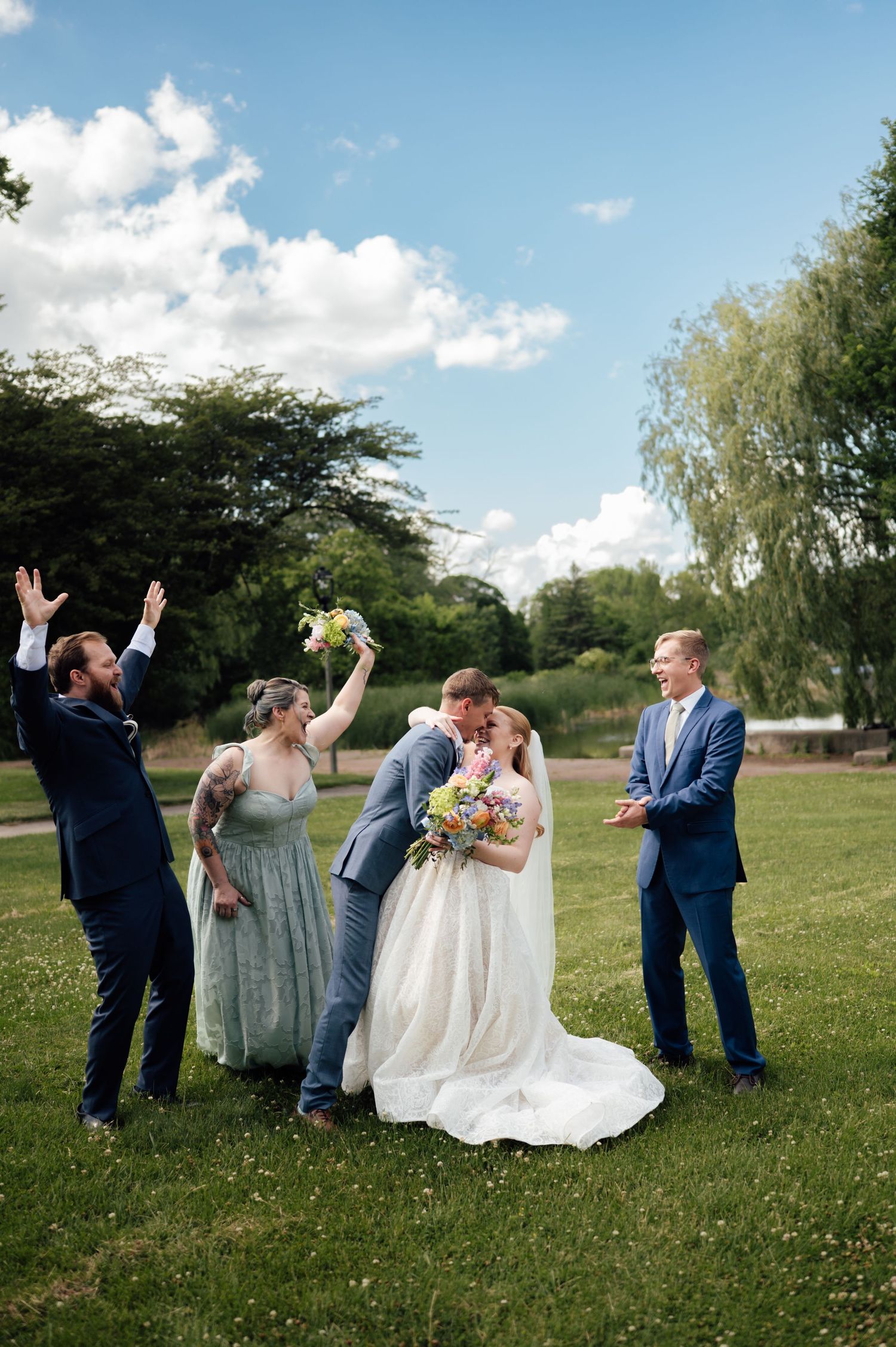 Bride and groom share a kiss outdoors while wedding party celebrates around them on a sunny green lawn.
