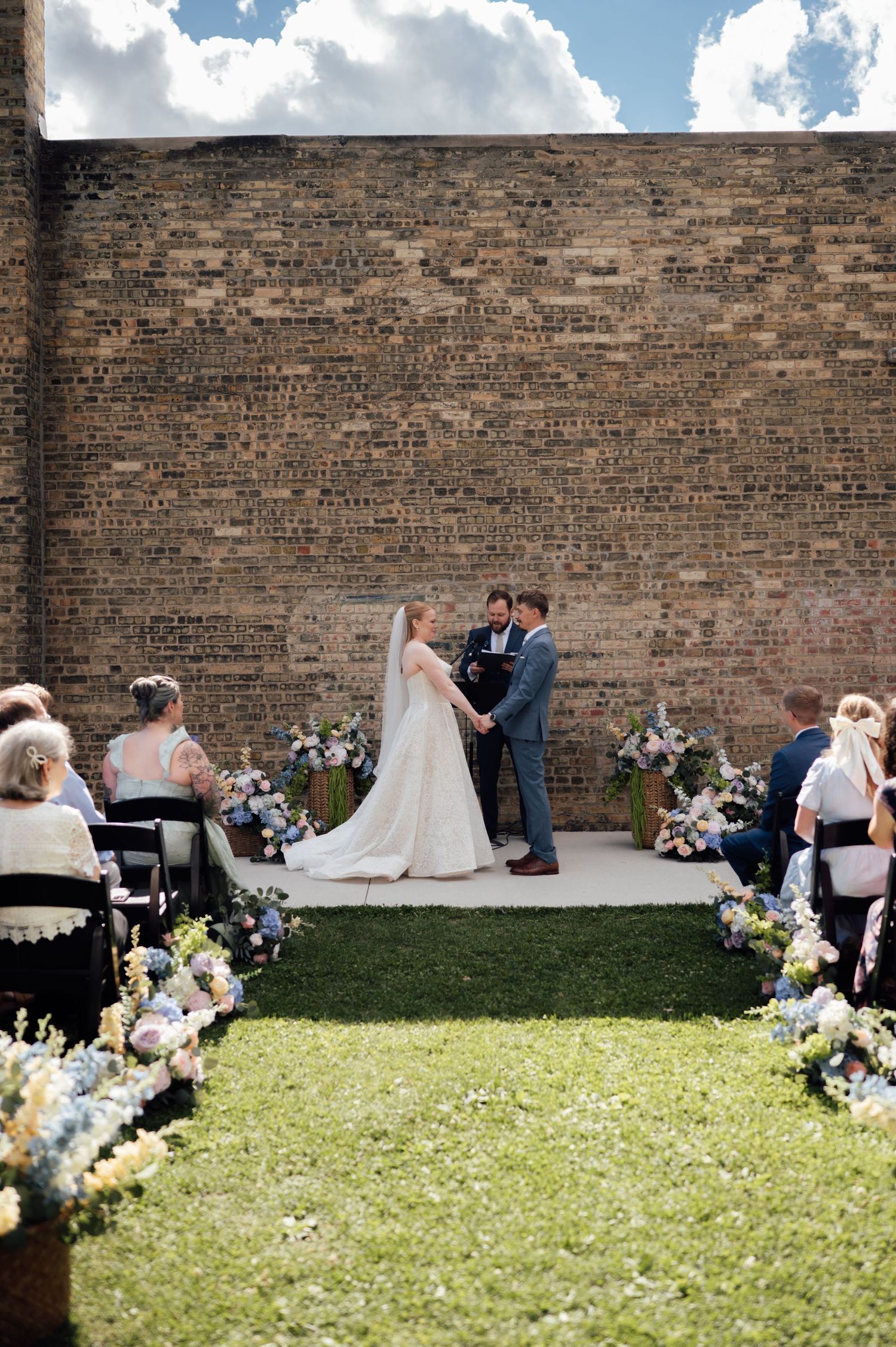 Outdoor wedding ceremony with bride and groom at brick wall altar, flanked by floral arrangements and seated guests on green lawn.
