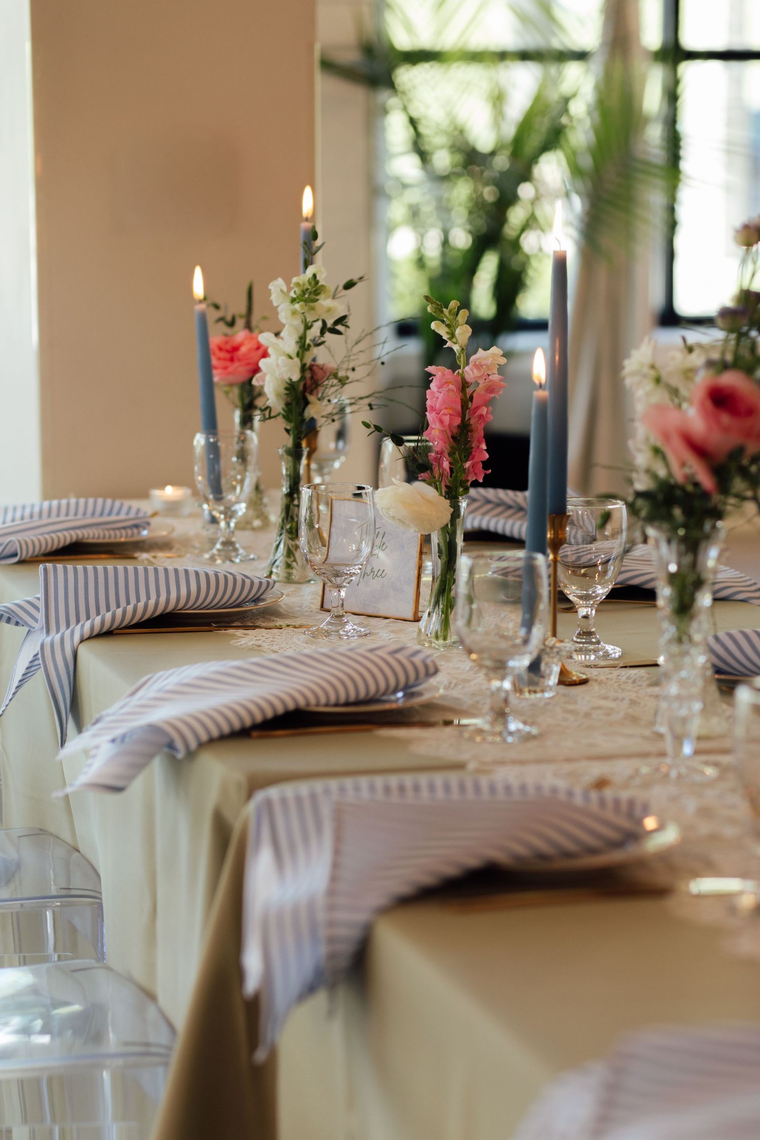 Elegant wedding table setting with blue candles, pink flowers, striped napkins, and glassware on a neutral tablecloth.