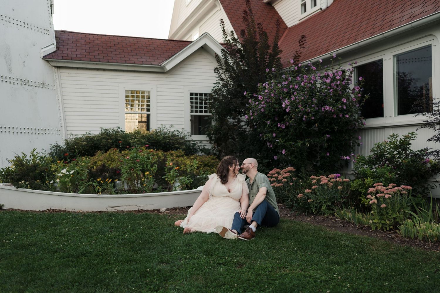 Couple sitting together on green lawn in front of historic white building with colorful flower gardens.