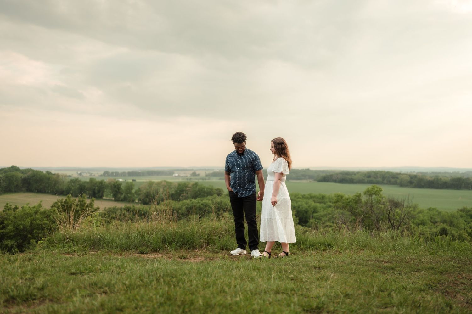 Couple standing on a scenic green hilltop overlooking a vast countryside landscape under cloudy skies.
