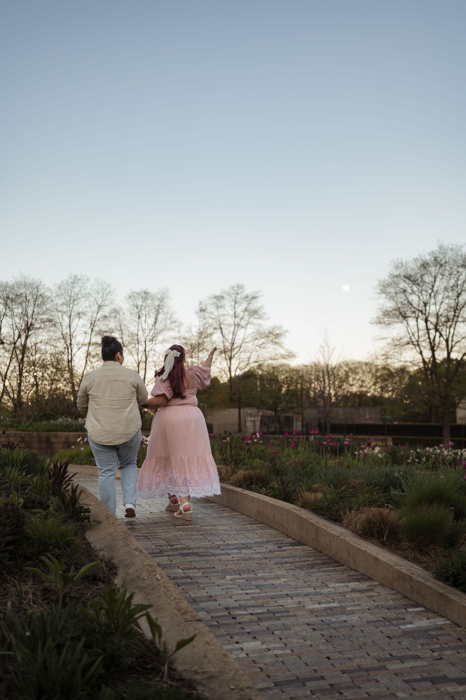 Couple walking hand in hand along a garden path at sunset pointing at moon.