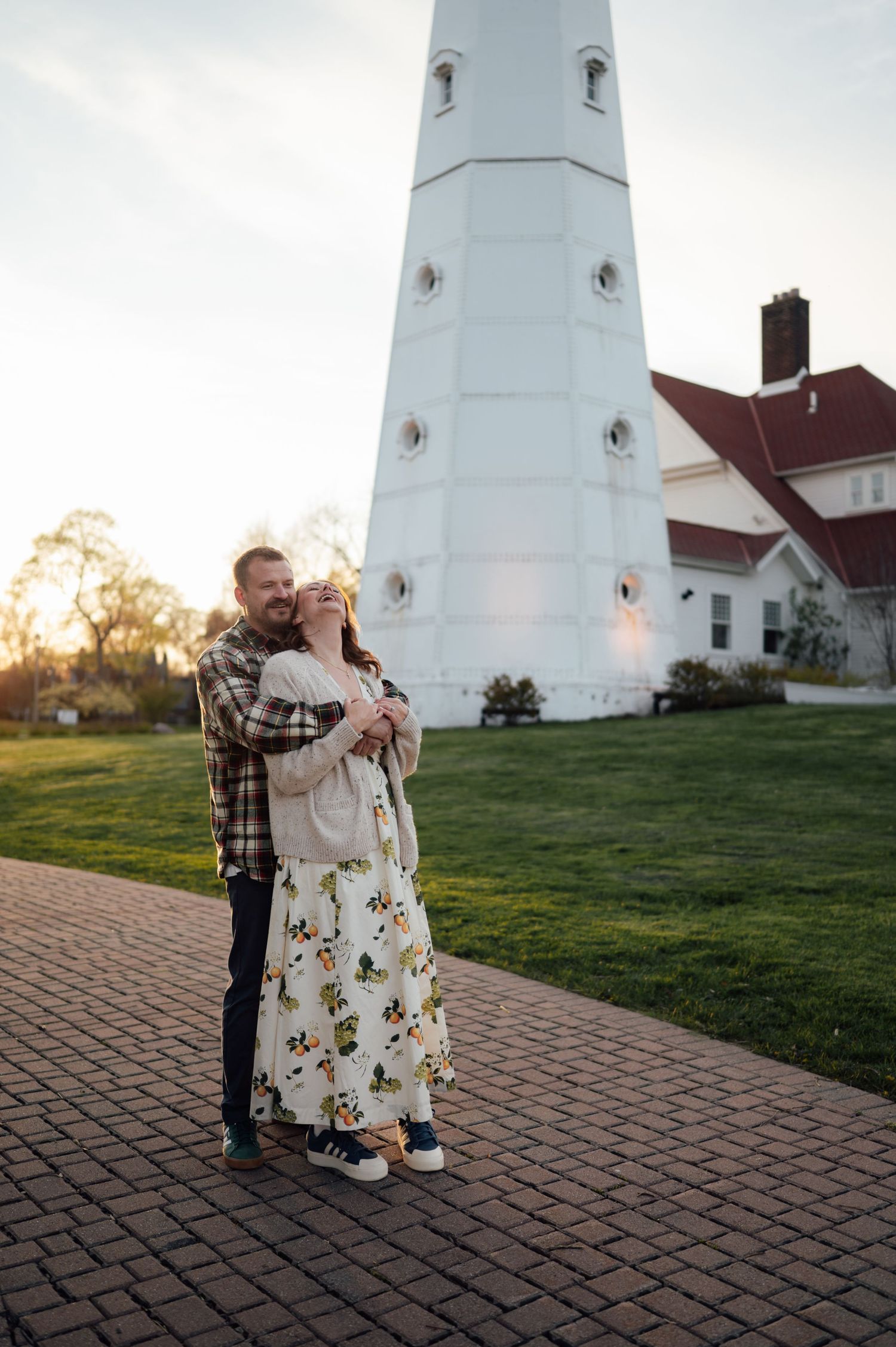Couple embracing at golden hour in front of a tall white lighthouse with a red-roofed keeper's house.
