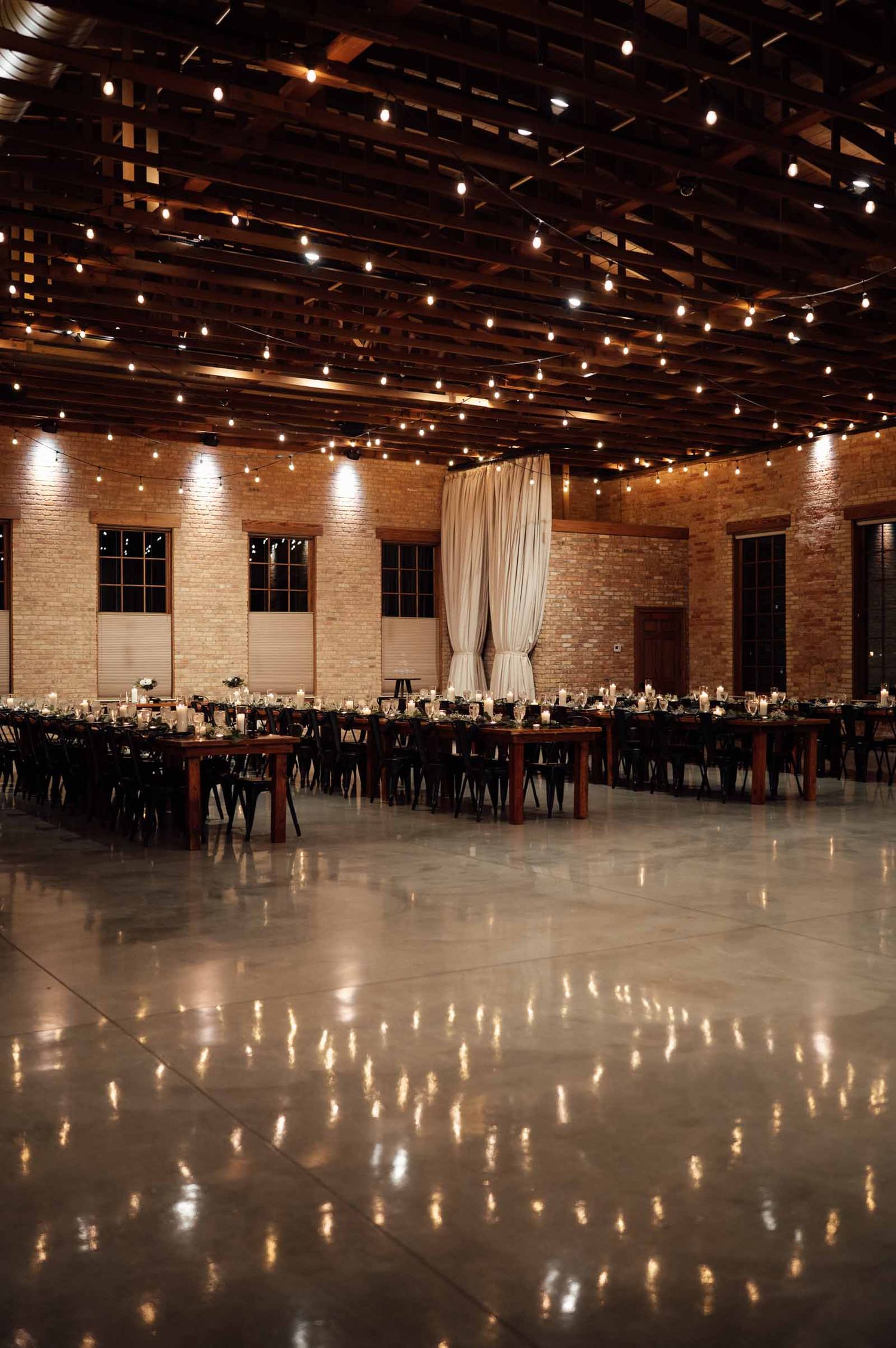 Elegant wedding reception hall with string lights, long dining tables, and brick walls in an industrial venue.