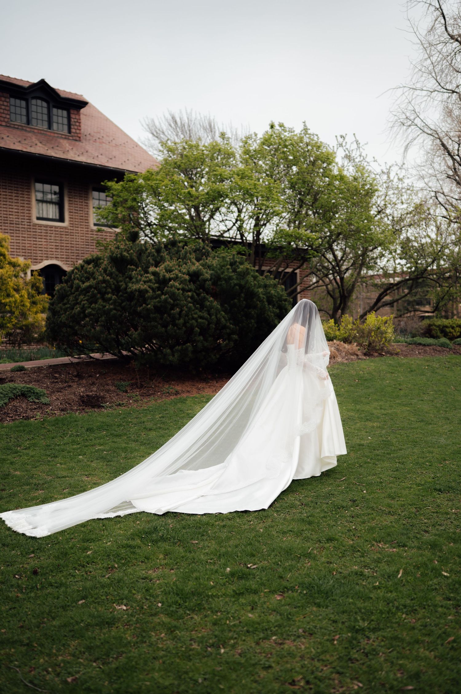 Bride in white gown with dramatic cathedral veil walks across lush green garden lawn.